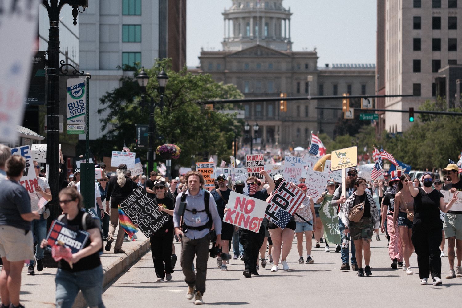 Protesters march on a street with signs and banners near government buildings.