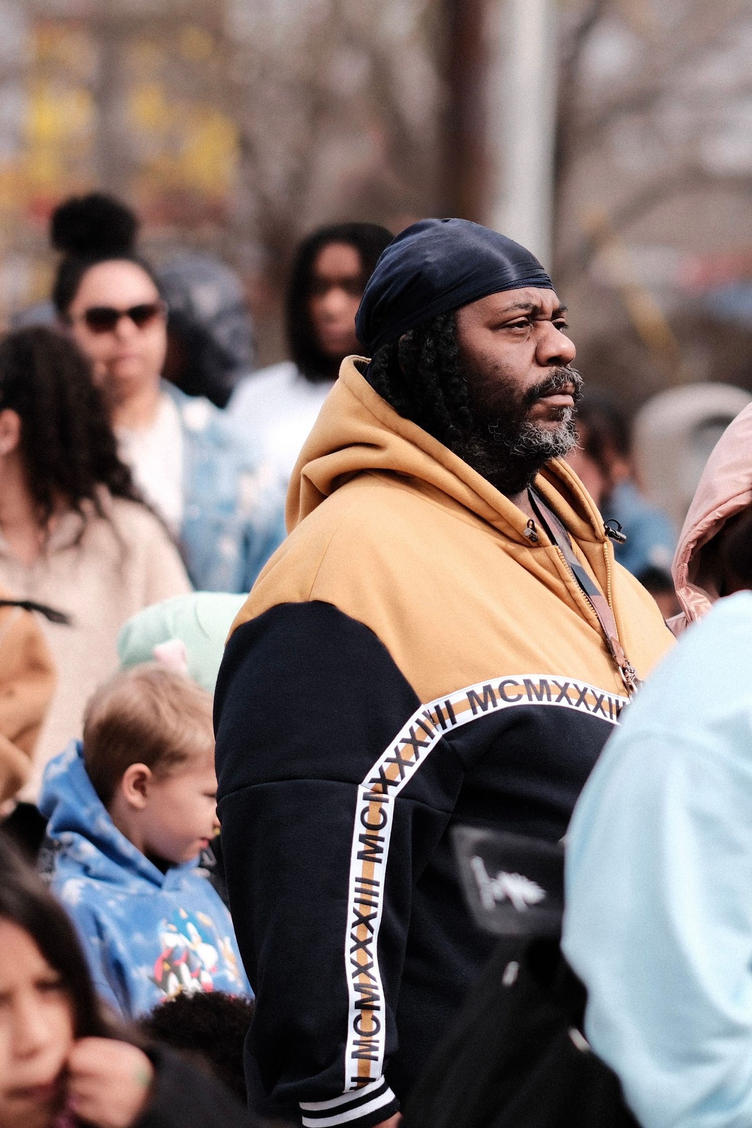 Person in camel-colored hoodie and black clothing at outdoor gathering among crowd of people.