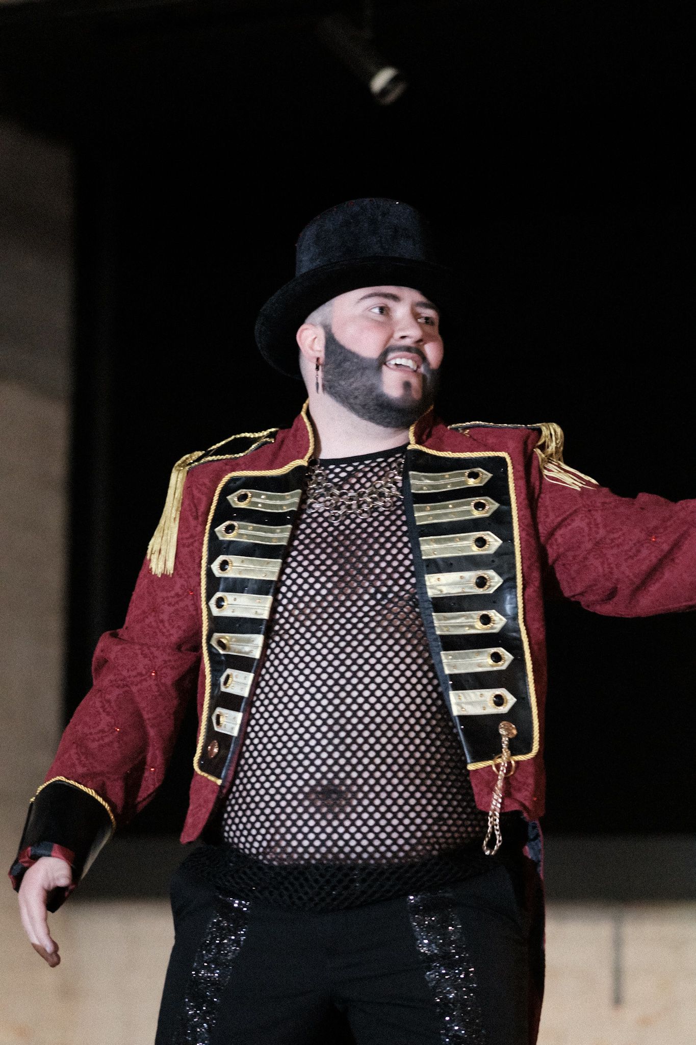 Performer wearing black hat and red military-style jacket performing on stage in dramatic lighting.