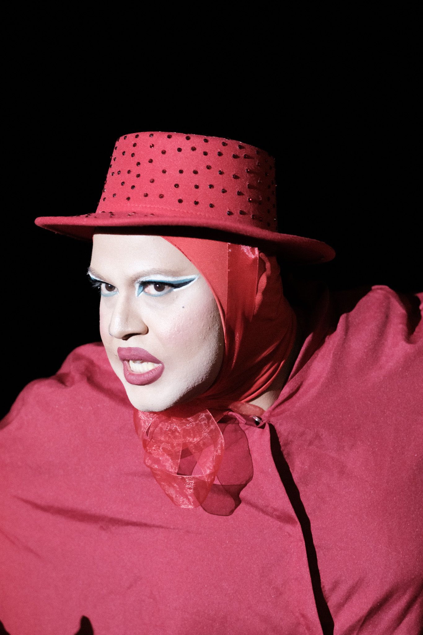 A dramatic portrait in deep red featuring studded hat, bold makeup and ruffled collar against a black background.