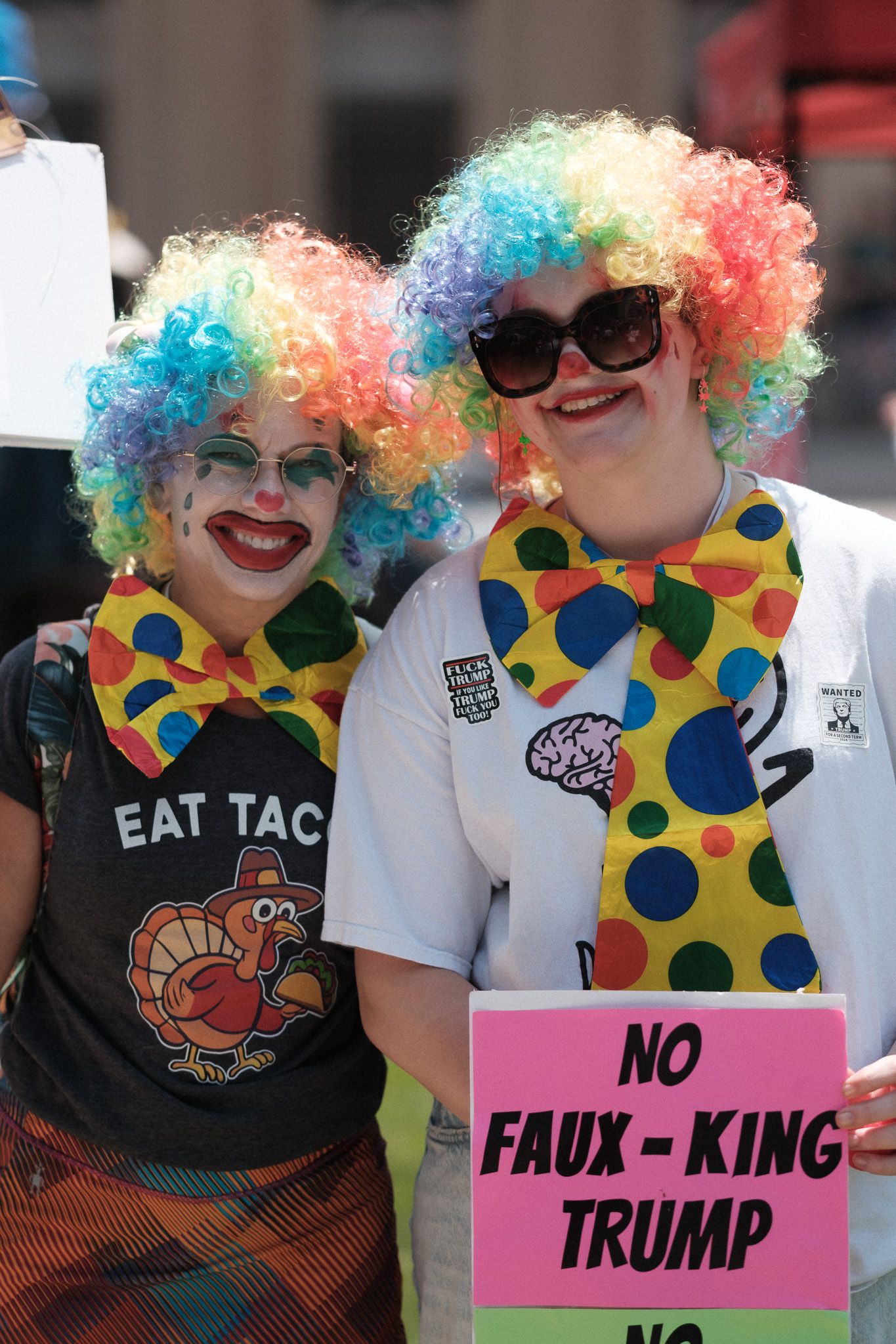 Protesters wearing colorful clown wigs and polka dot outfits hold pink signs during a demonstration.