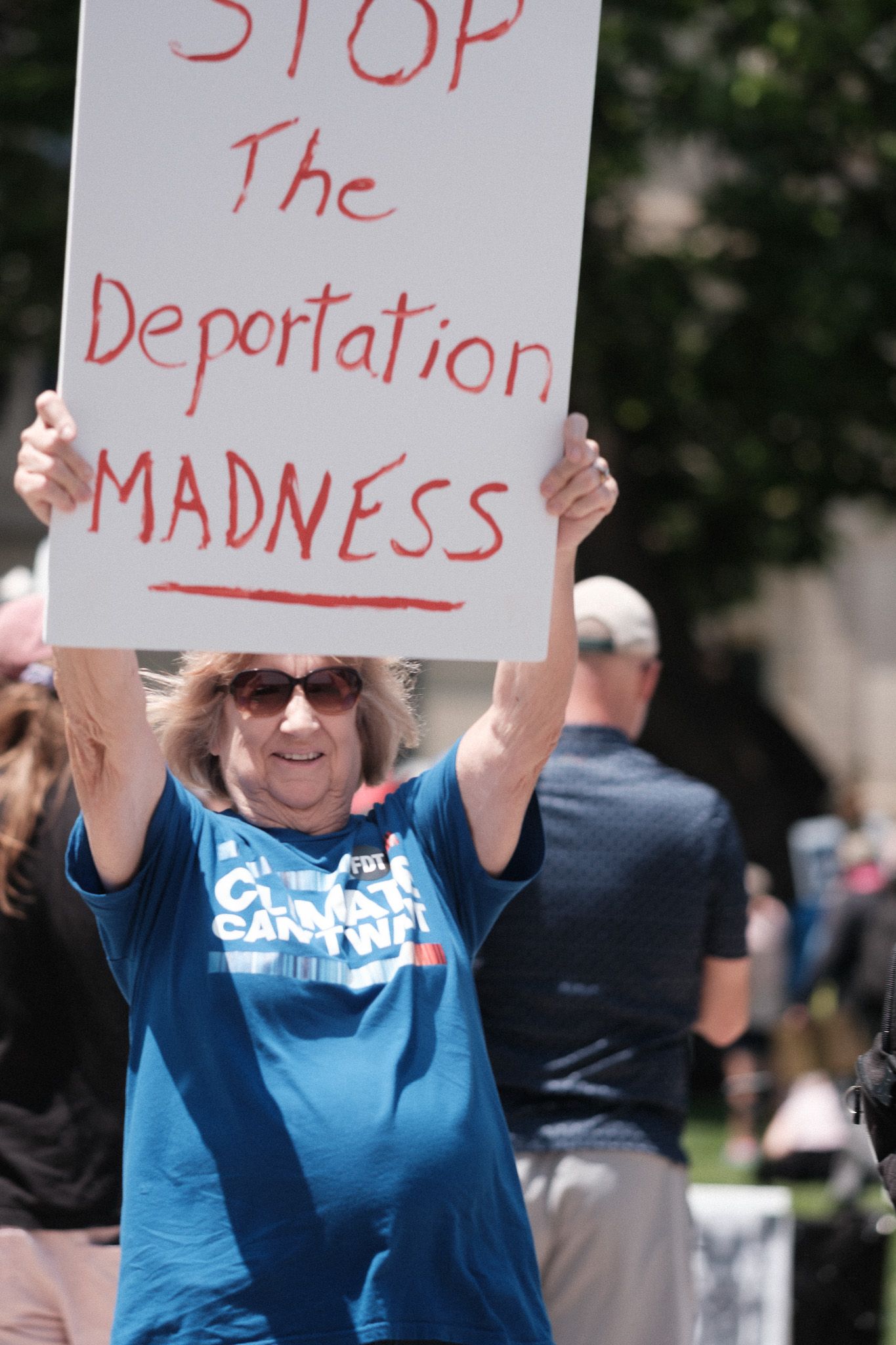 A protester in a blue t-shirt holds up a red handwritten sign about deportation.