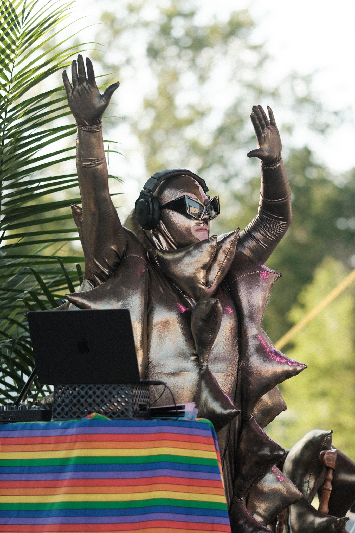 DJ in shiny costume and headphones performs behind rainbow-striped table at outdoor festival.