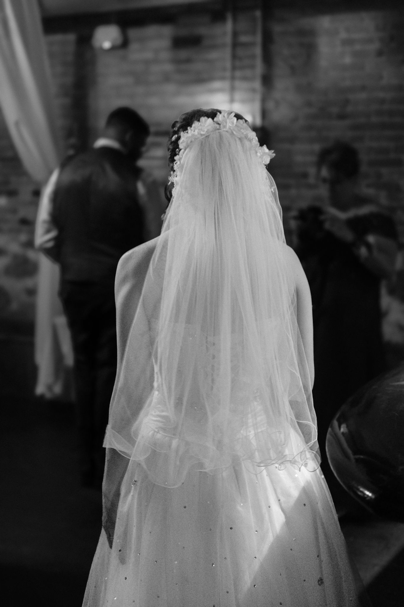 A bride wearing a white wedding gown and long flowing veil stands in an indoor venue with exposed brick walls.