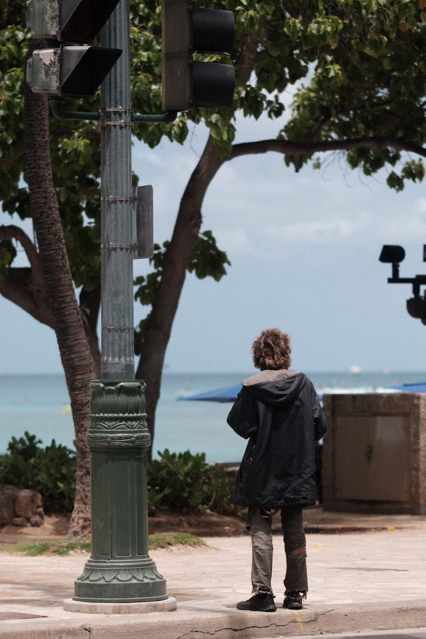 Someone in a dark hoodie stands alone by a vintage lamppost overlooking the ocean on a cloudy day.