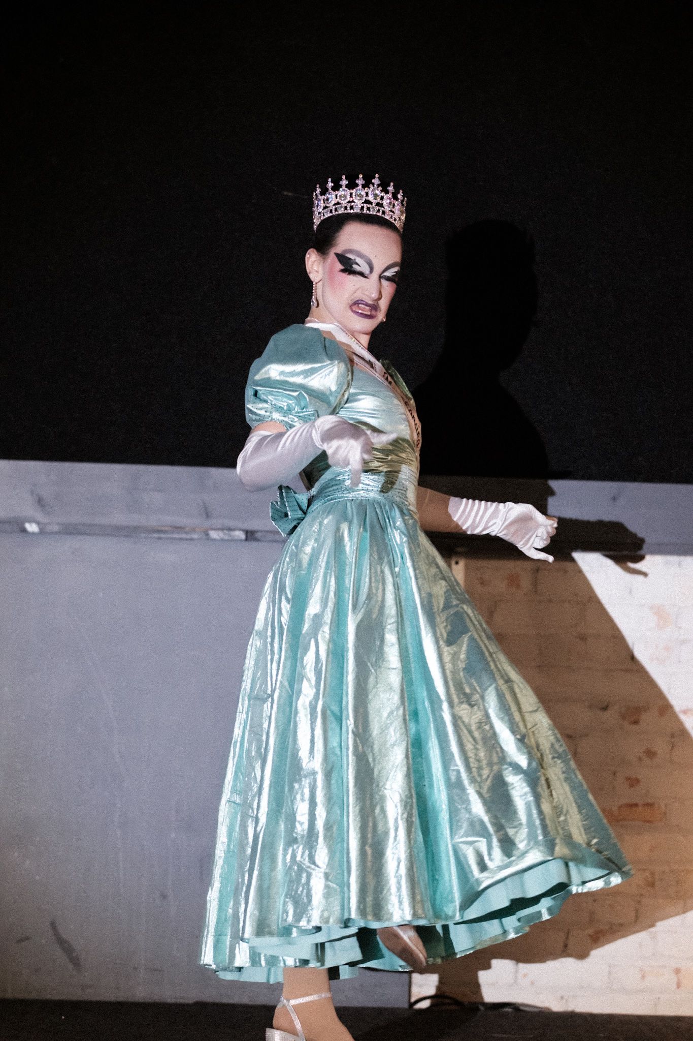 Performer in mint green vintage dress and crown strikes theatrical pose on stage.