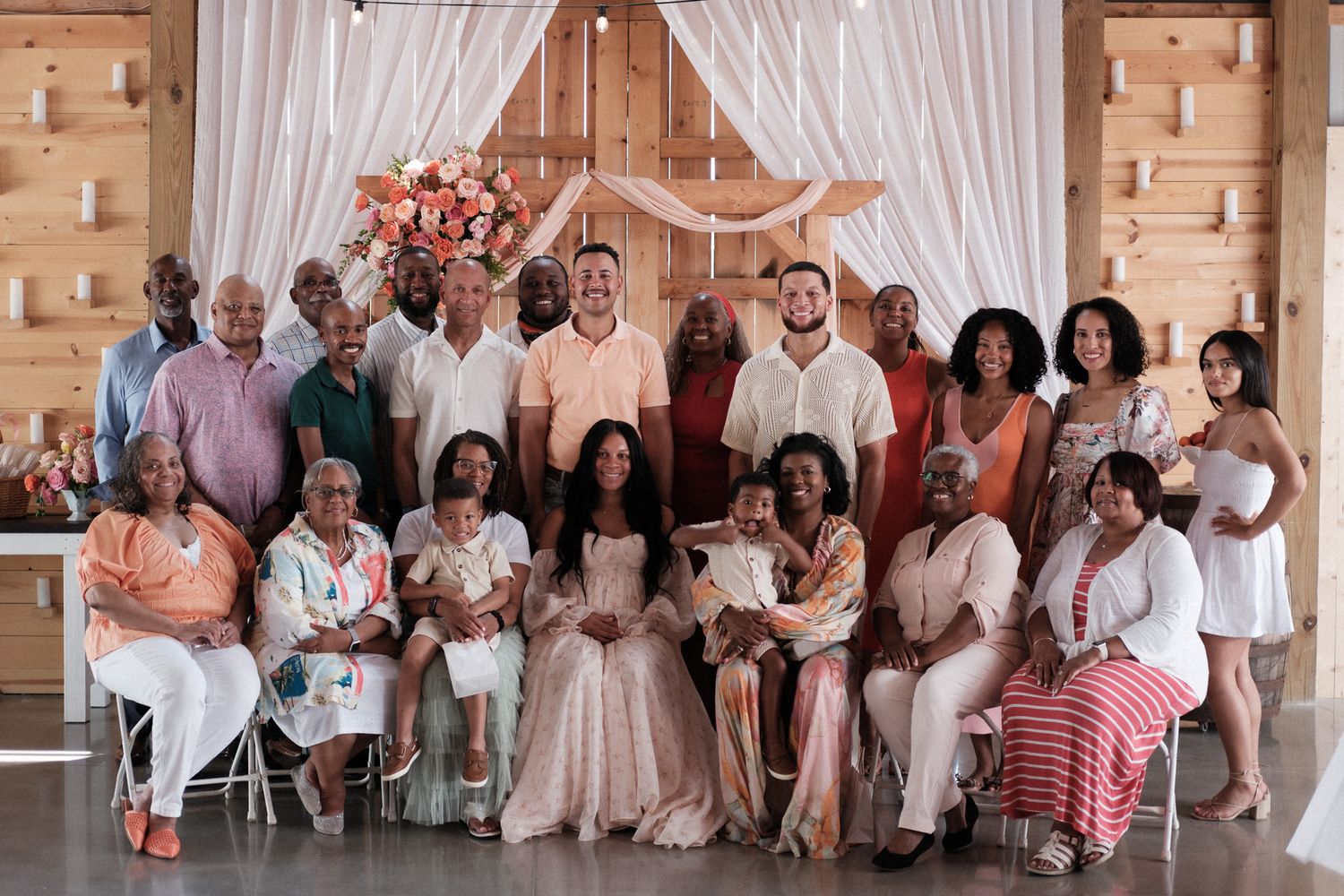 A large group of people gathered in an elegant barn venue decorated with pink floral accents for a celebratory portrait.