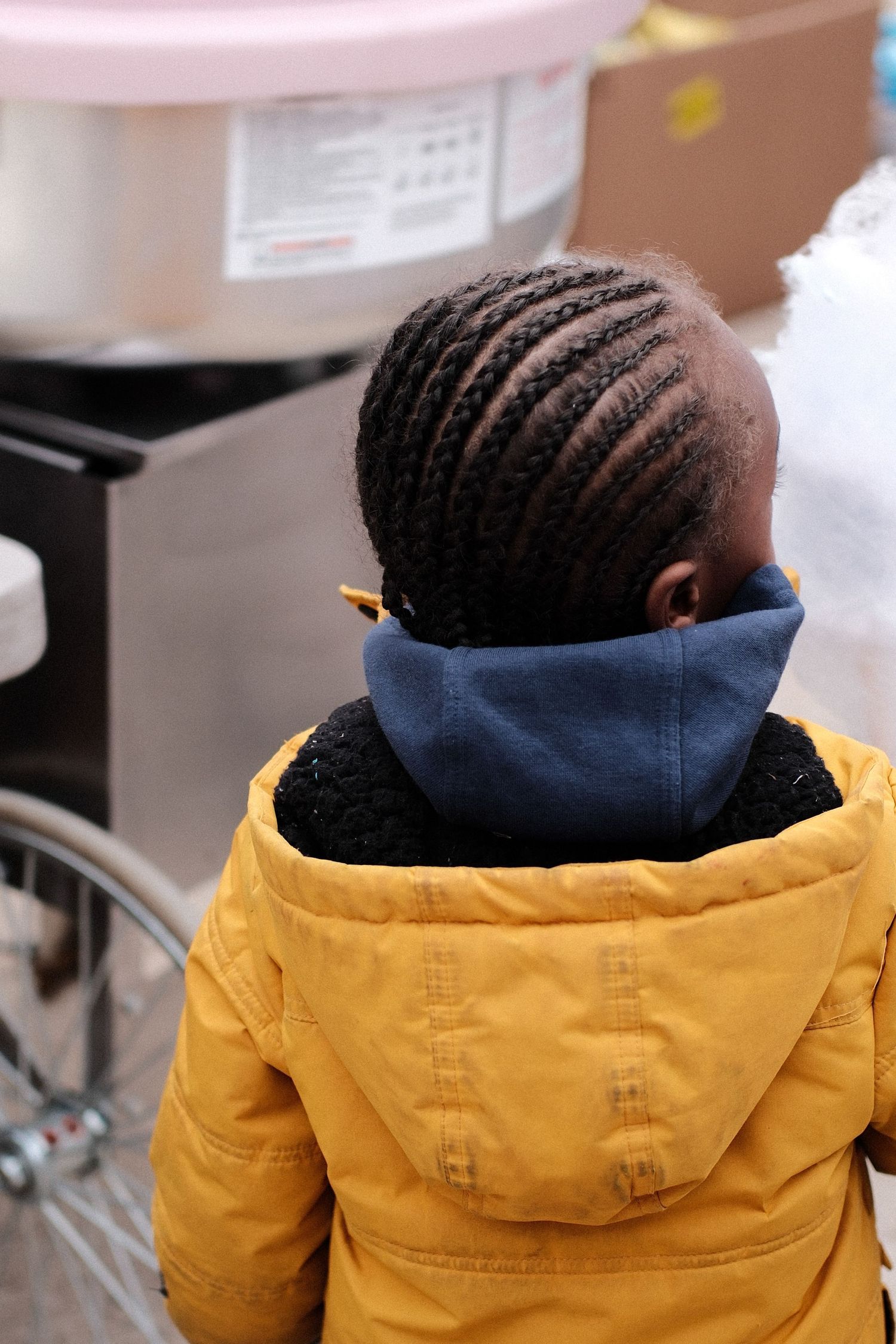 Back view of child wearing yellow winter coat and blue scarf with neatly braided cornrow hairstyle.