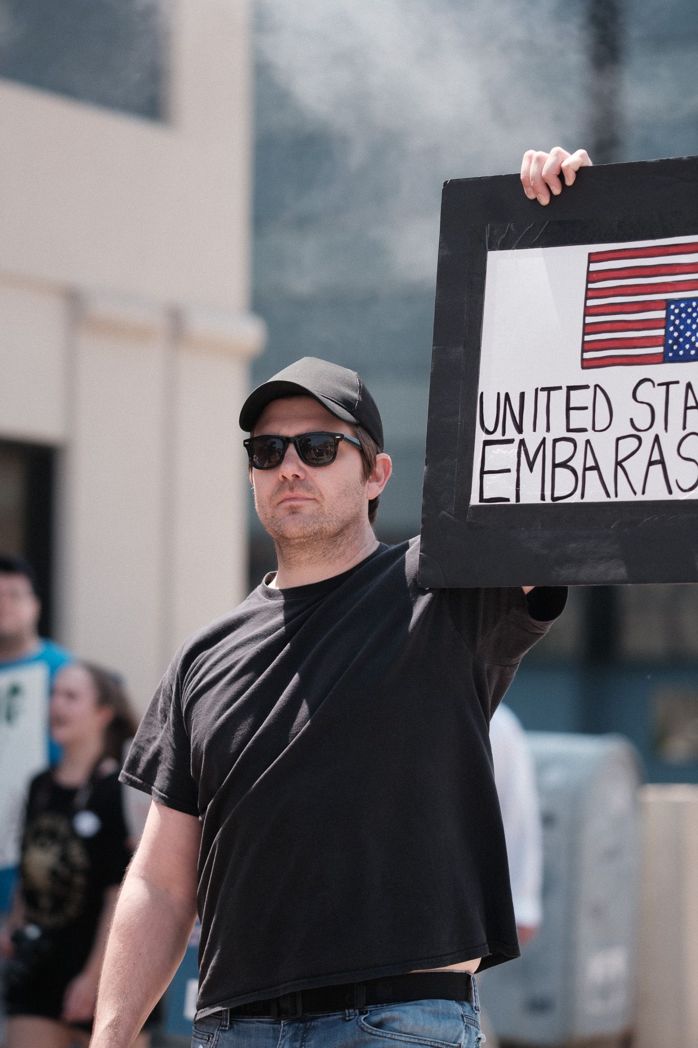 Person in black shirt and cap holds protest sign with American flag at outdoor demonstration.