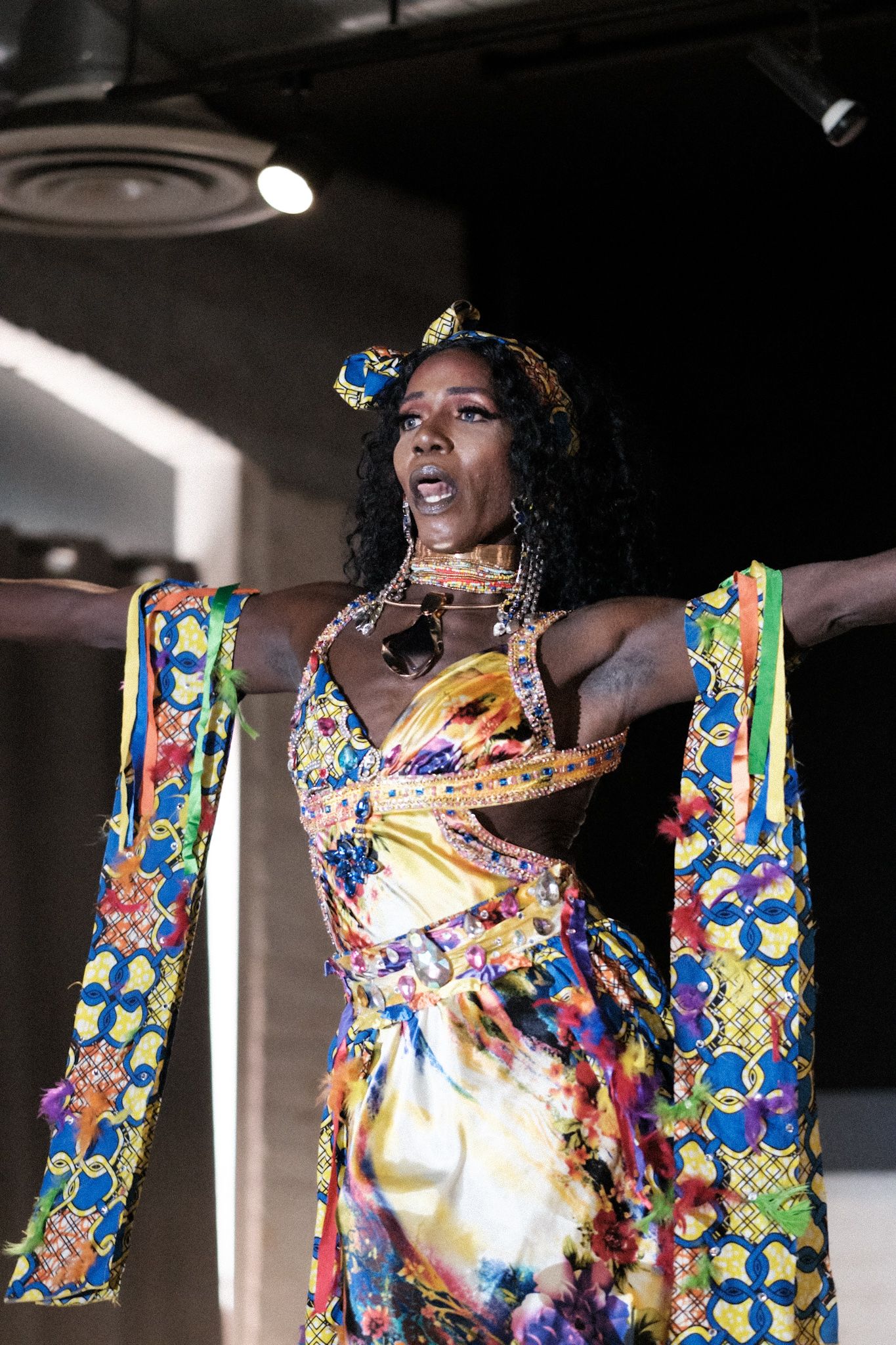 Performer in colorful floral print dress with dramatic sleeves poses expressively on stage.