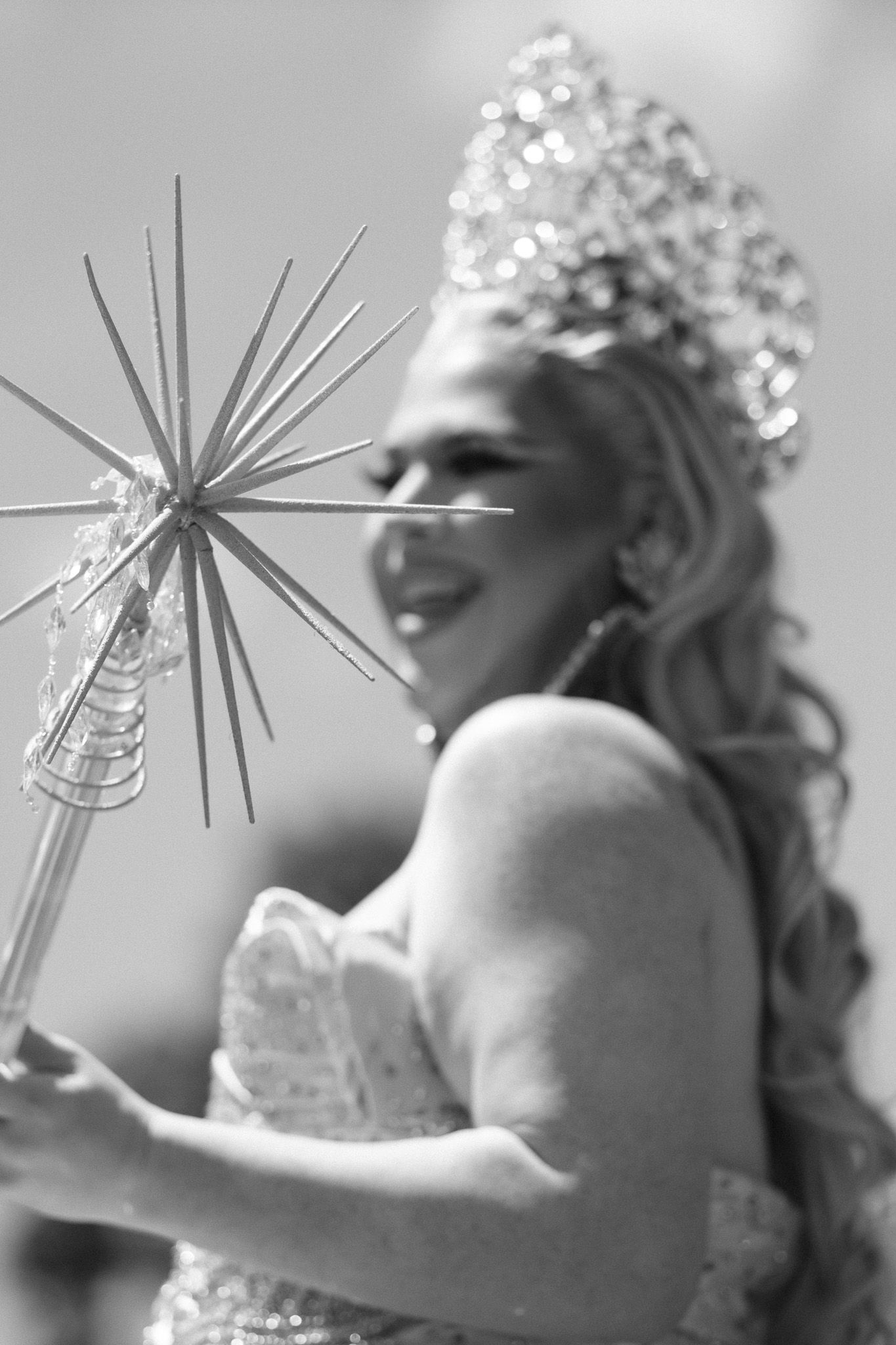 A performer wearing a sparkly crown and white dress holds a star-tipped wand in this black and white photo.