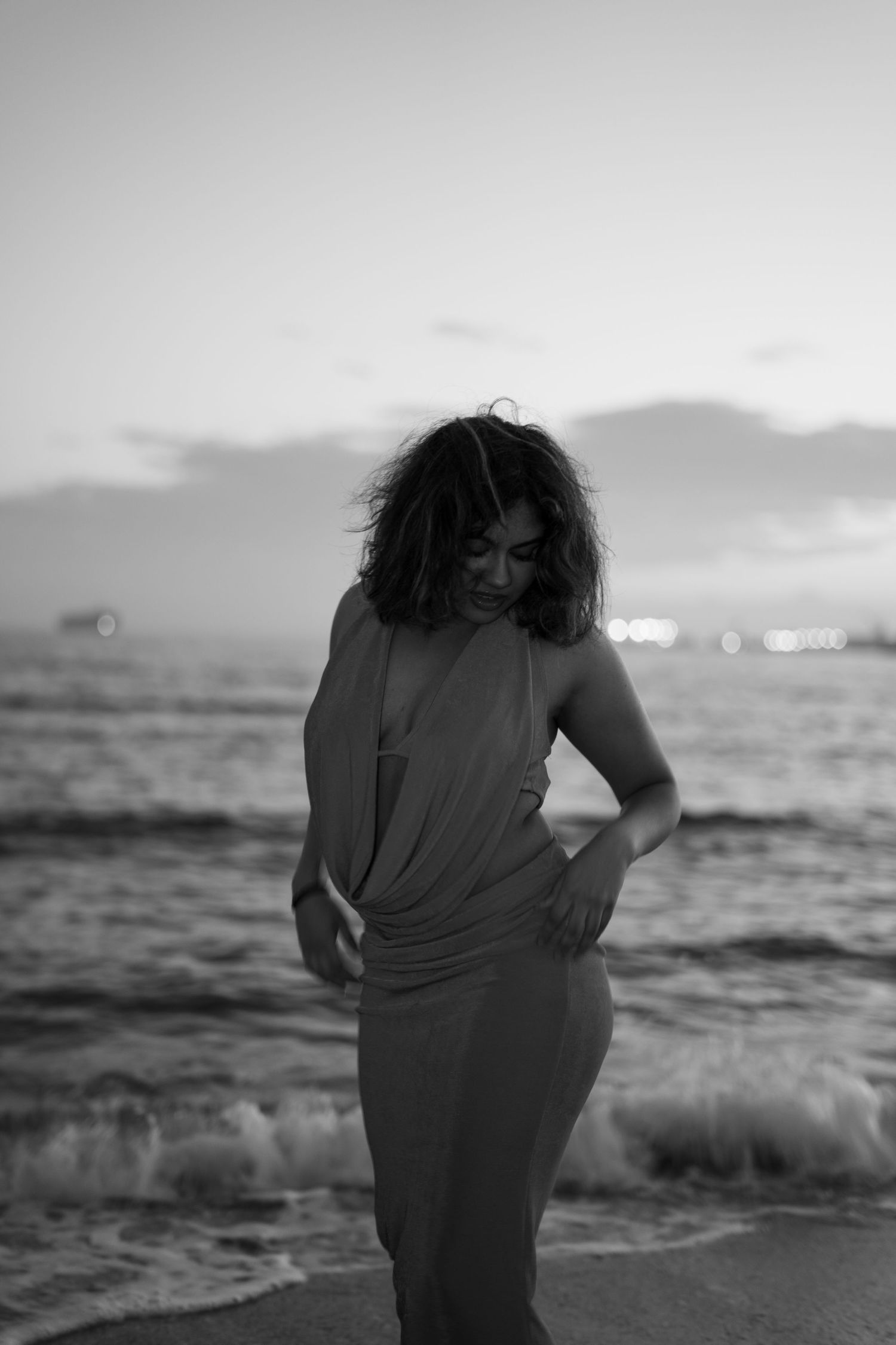 Grayscale photo of figure by ocean waves during sunset.