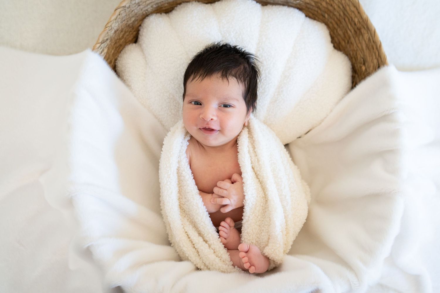 A newborn baby wrapped in a soft white blanket smiles while lying on white bedding.