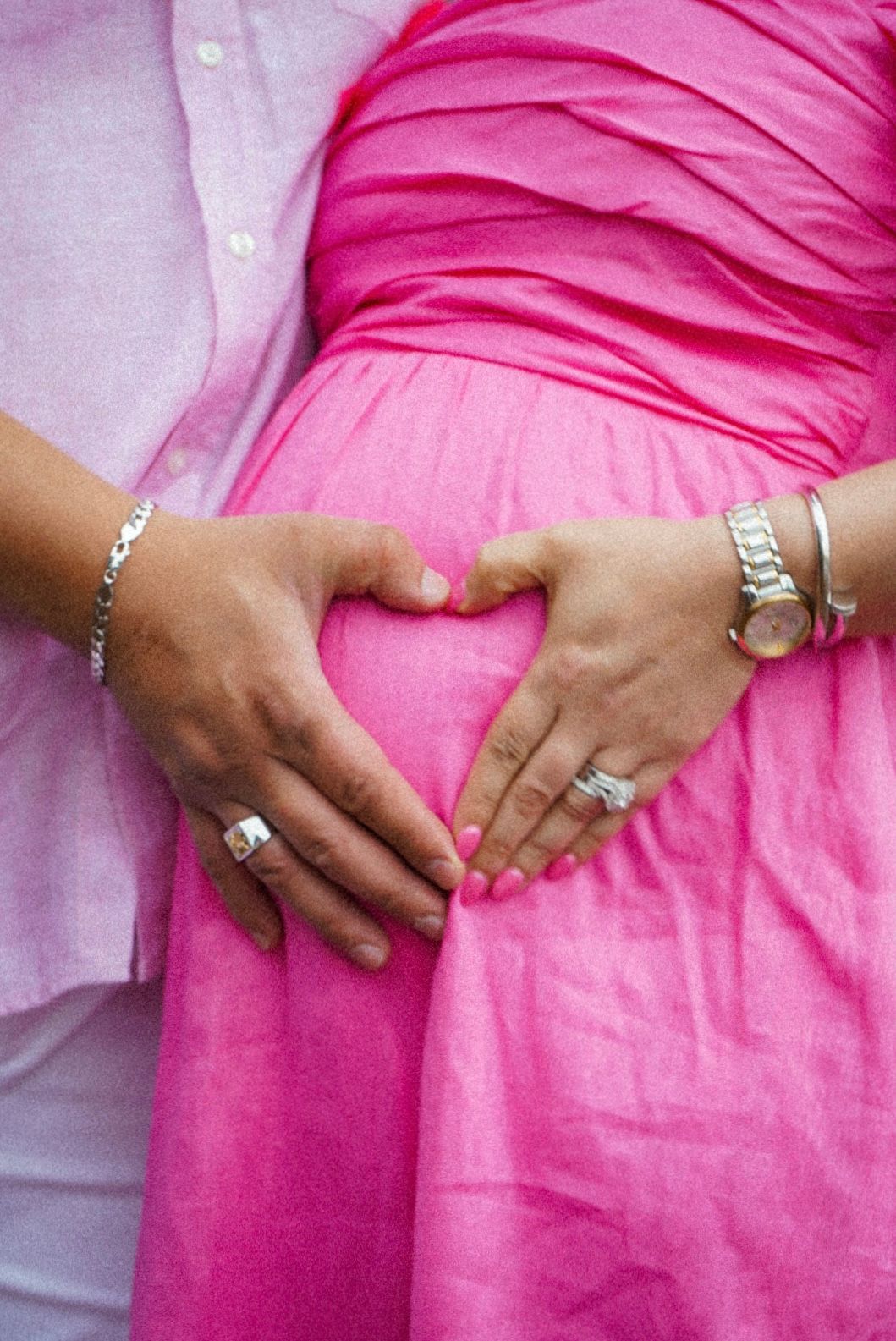 Two hands creating heart shape against pink fabric with jewelry visible.