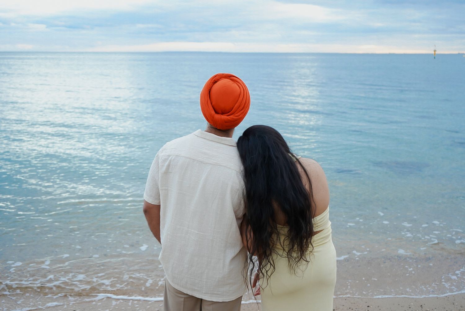 A couple walks along the shoreline at sunset, their silhouettes reflected in the calm ocean waters.