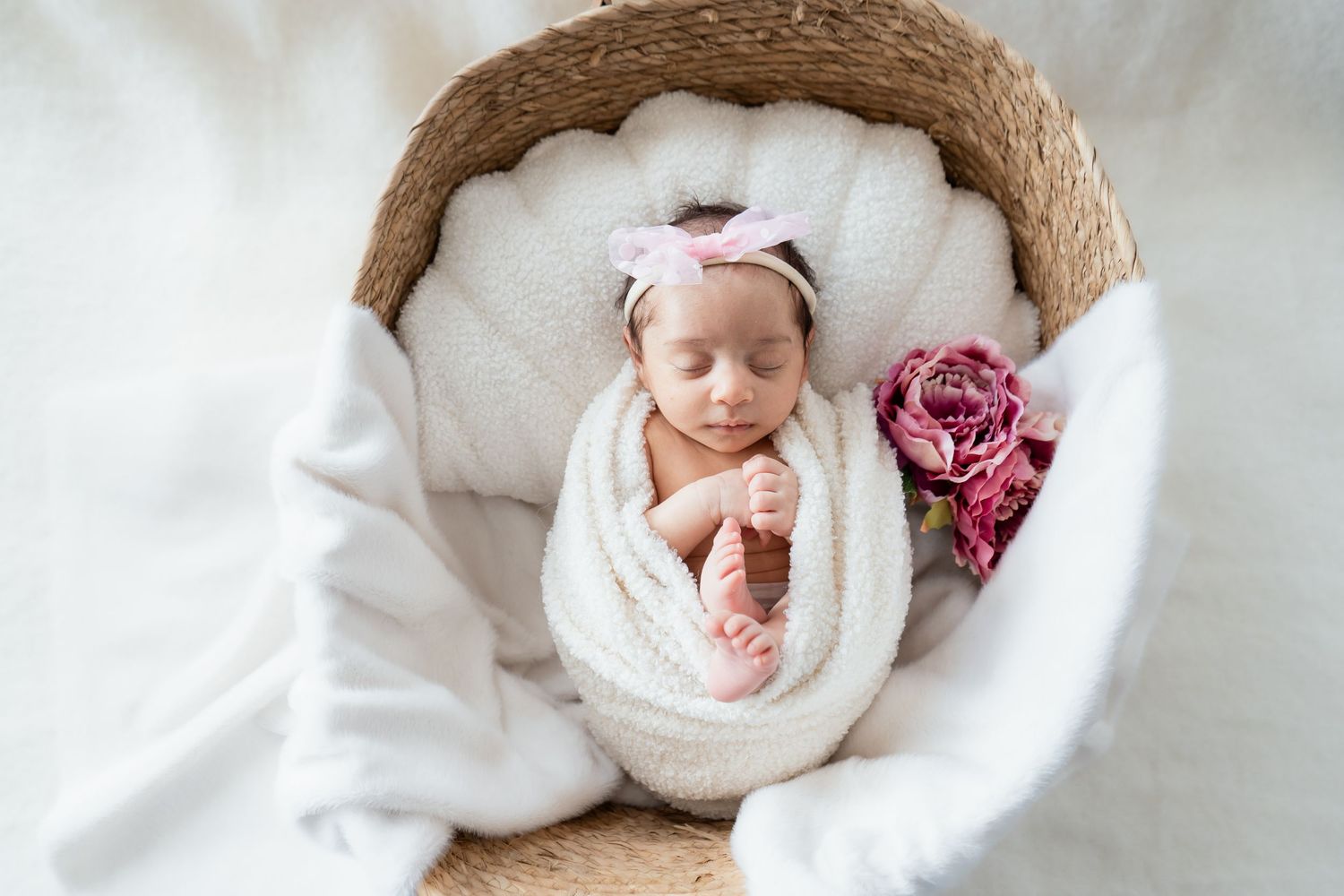 Newborn baby wearing white swaddle wrap sleeps peacefully on white bedding with pink floral accents.