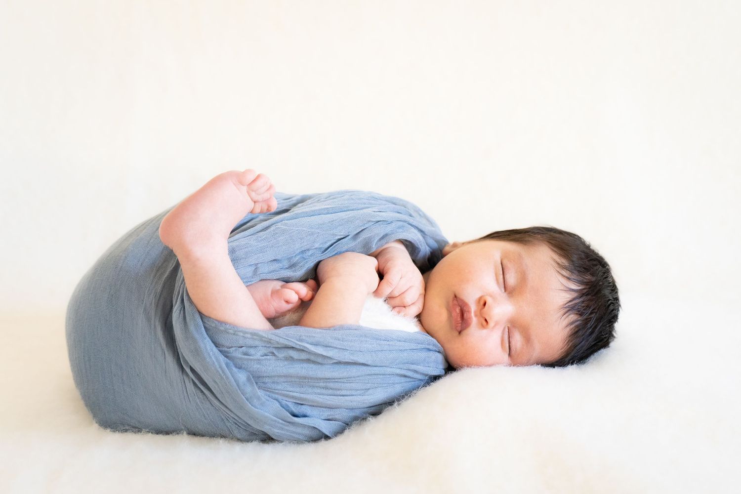 Newborn baby in light blue swaddle sleeps peacefully on white blanket in three similar poses.