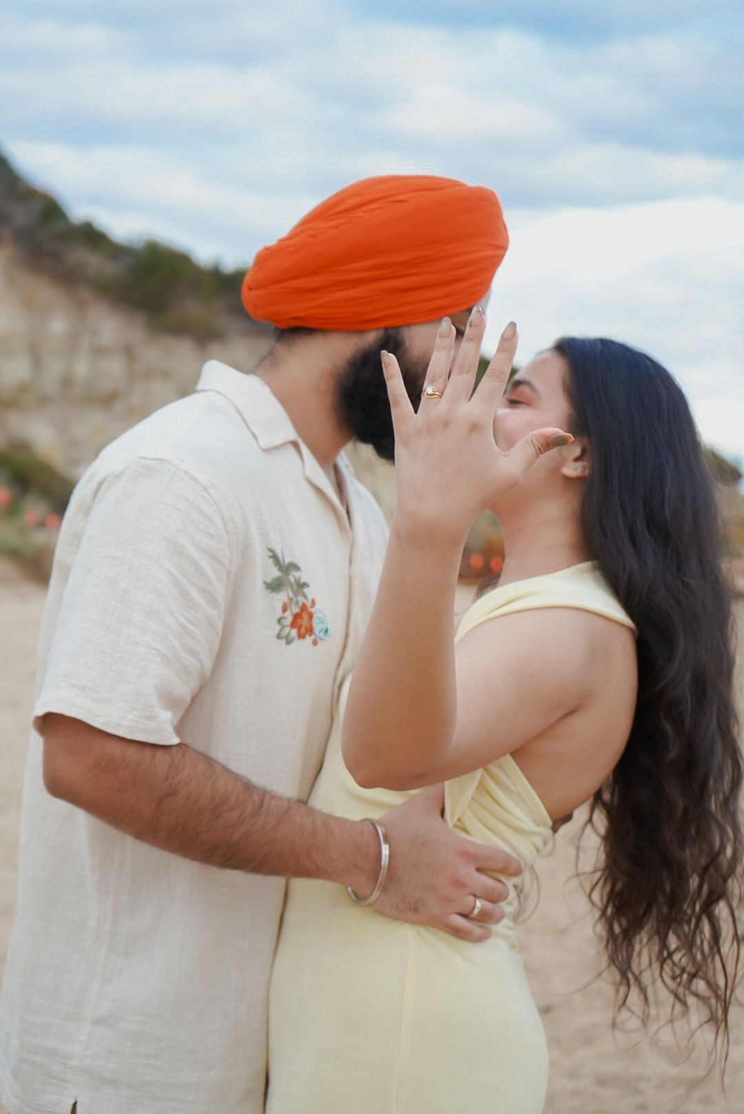 A couple shares an intimate moment on a sandy beach, with the woman in yellow and the man wearing orange headwear.