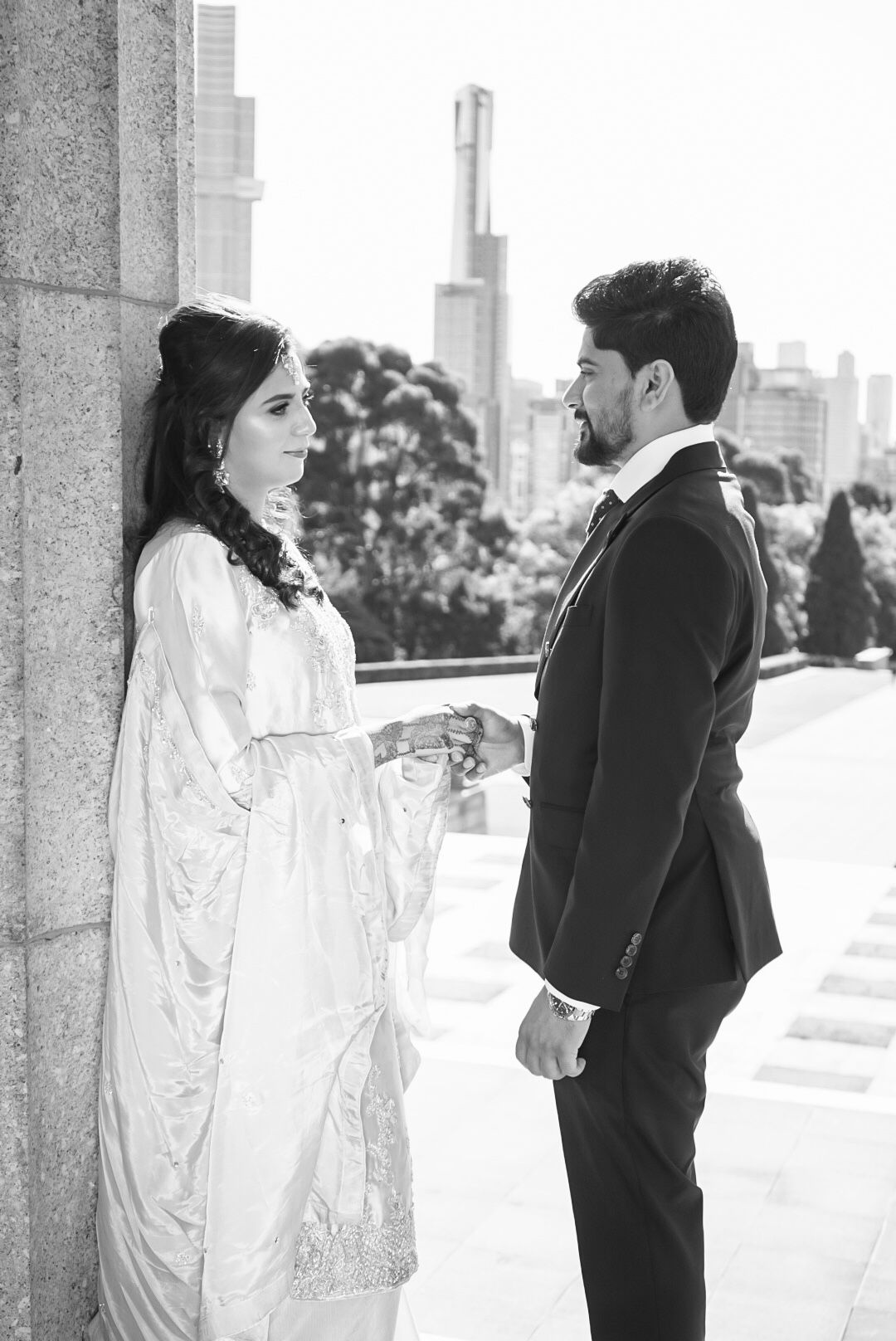 A couple in formal attire poses against a city backdrop in black and white.