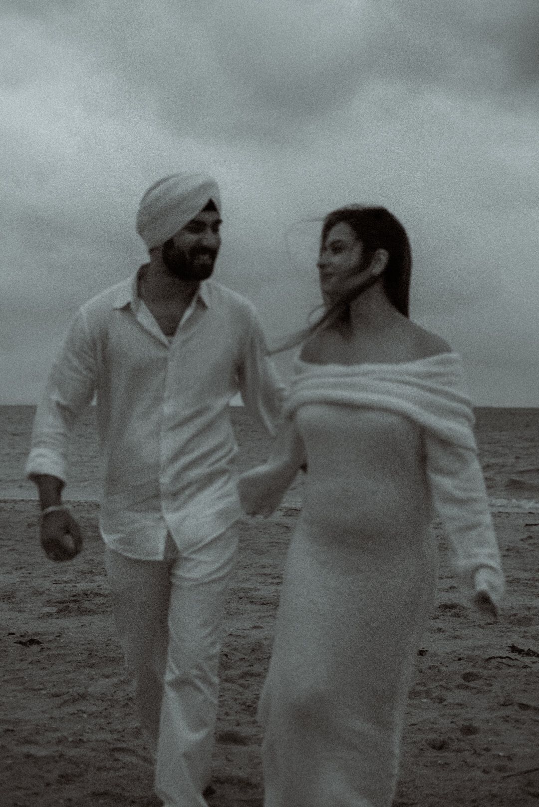 Black and white photo of couple walking on beach in white outfits.
