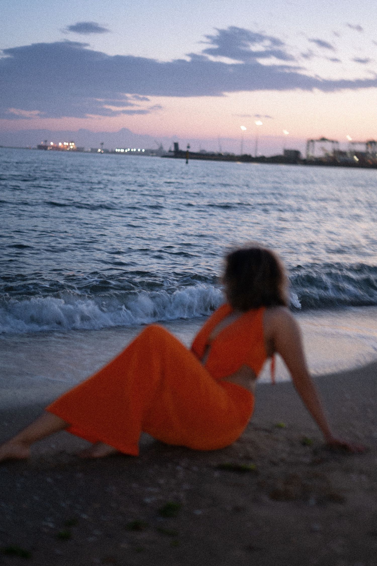 Silhouette in orange dress reclines on beach at sunset with ocean waves in background.