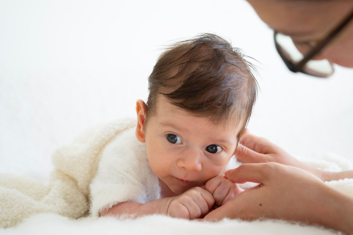 Curious newborn baby with blue eyes lying on soft blanket while parent gently holds tiny hand.