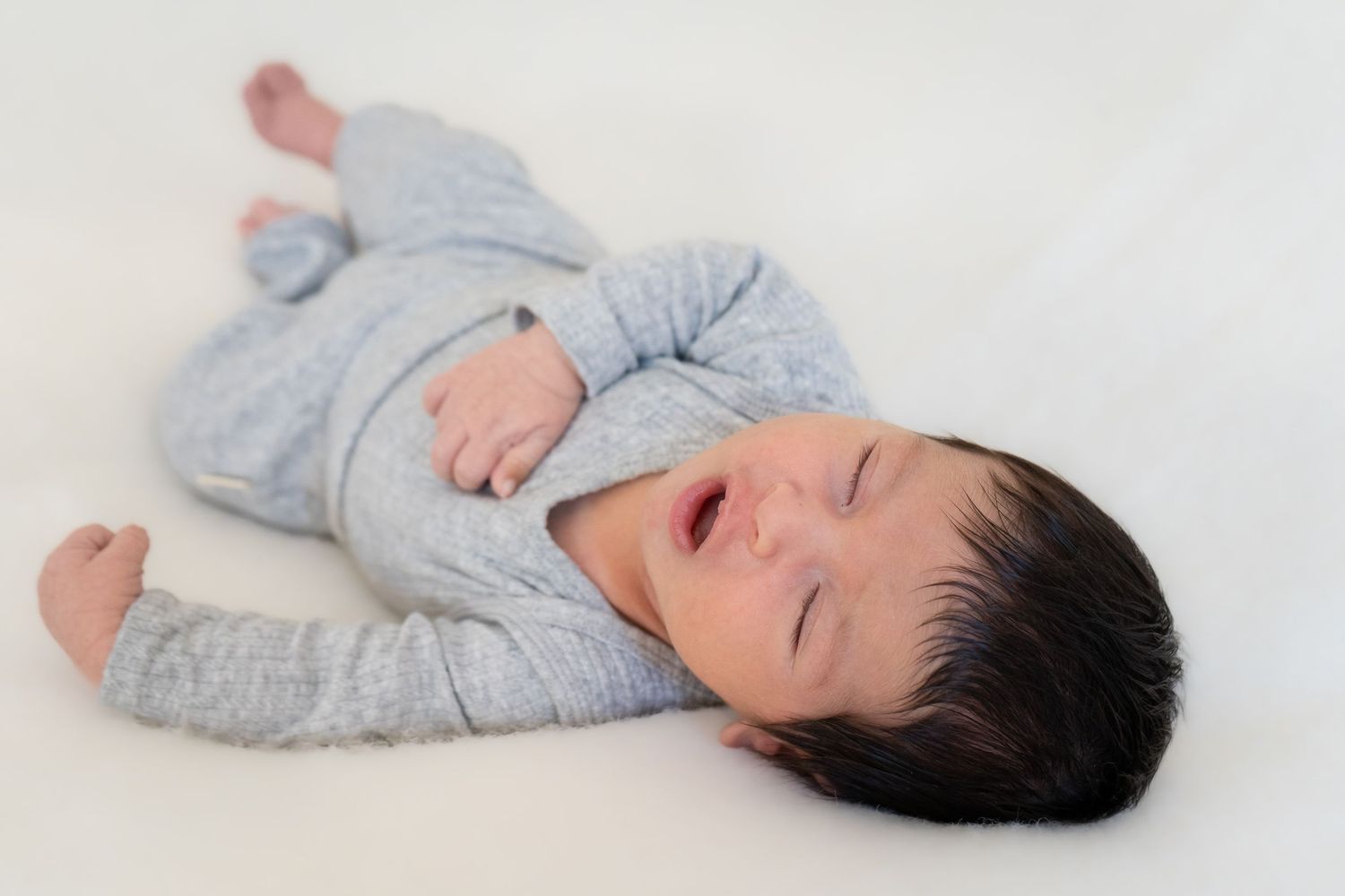 Baby wearing light blue sleepwear lies on white background while yawning.