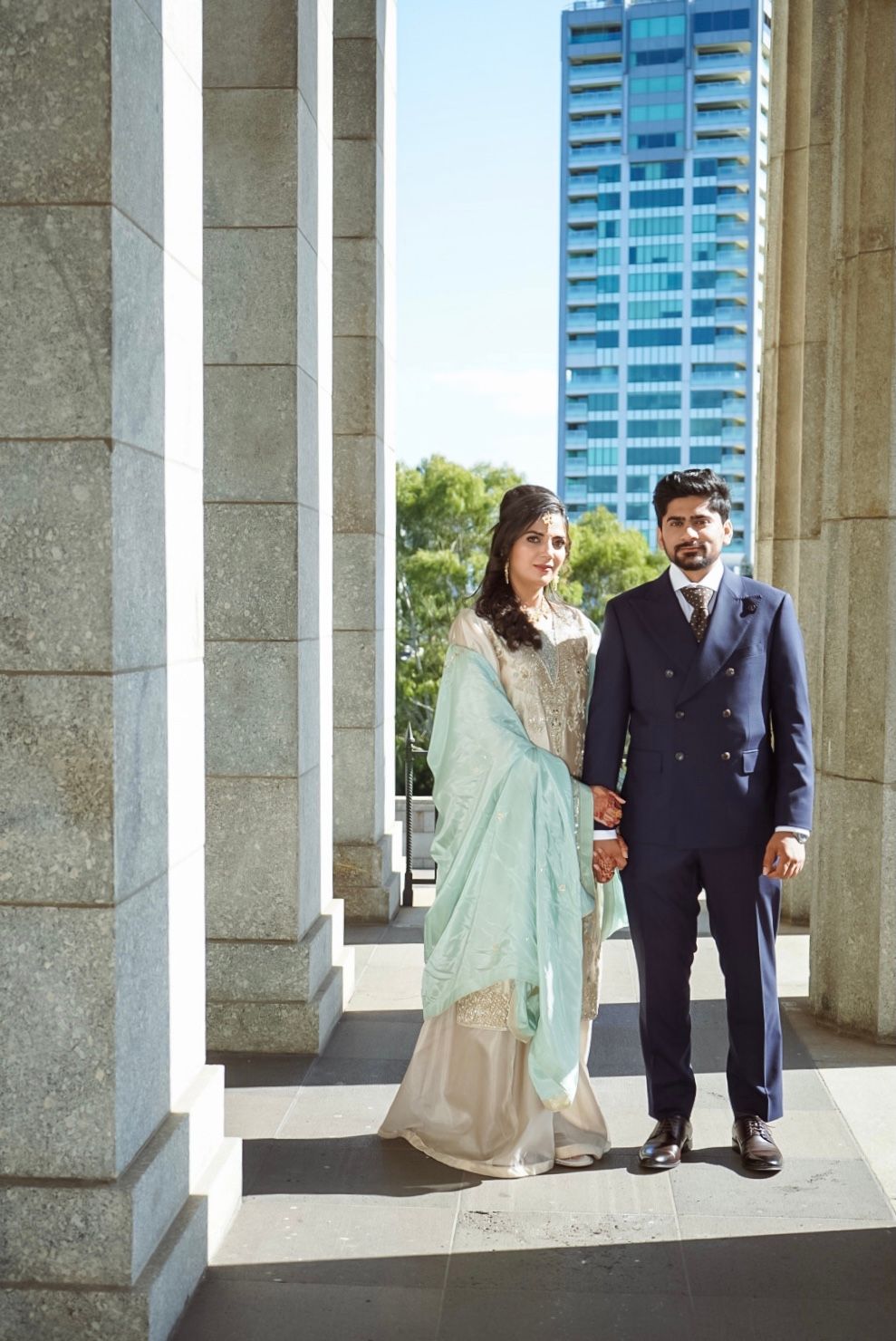 A couple in traditional wedding attire stands between elegant stone columns.