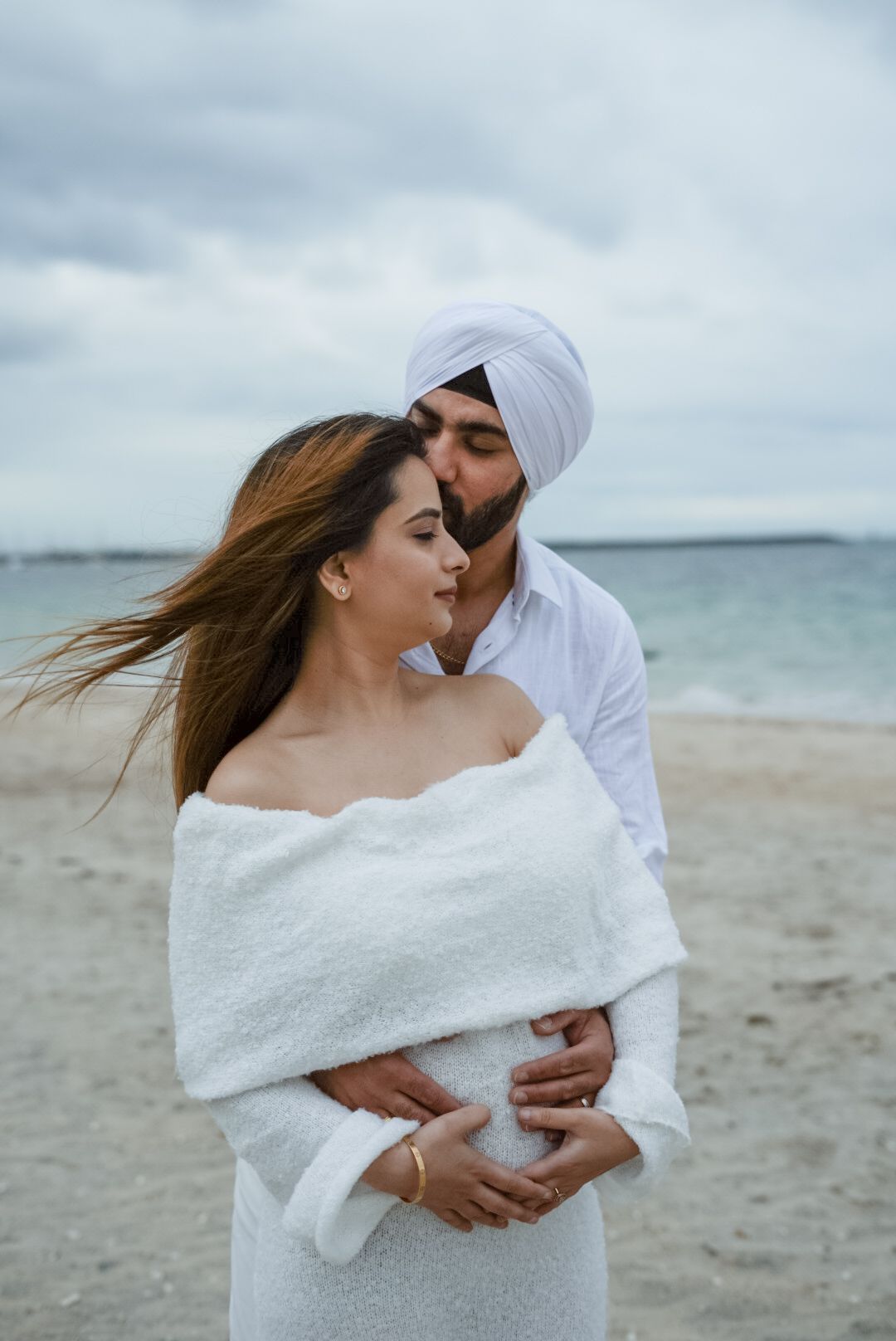 Couple embracing on beach with white clothing against cloudy sky.