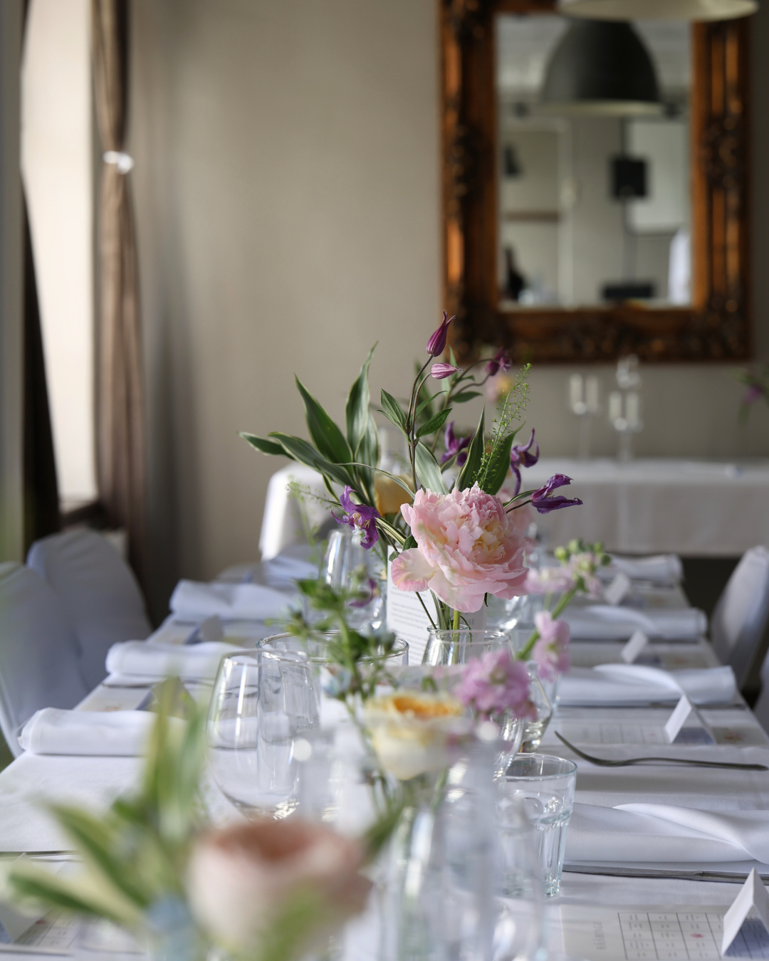 Elegant dining table set with white linens, glassware, and fresh floral centerpieces featuring pink peonies and purple blooms.