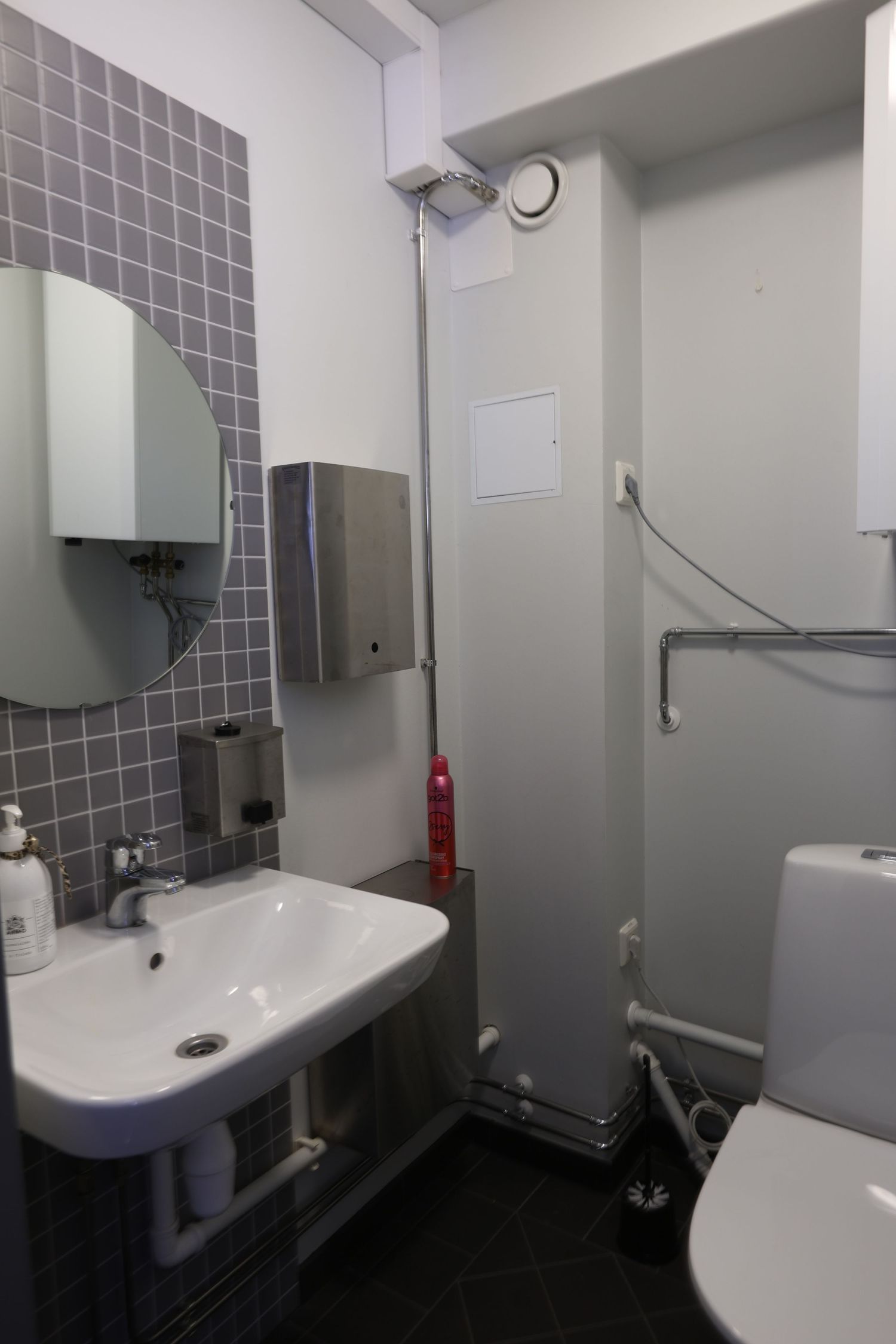 Compact bathroom with white sink, toilet, gray mosaic tiles, mirror, soap dispenser, and fire extinguisher.