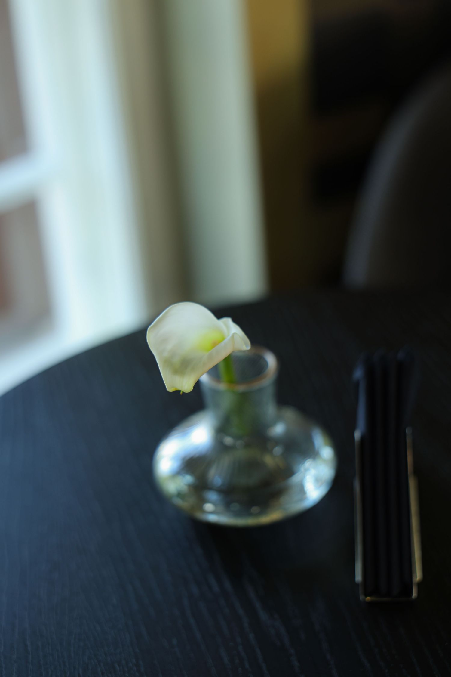 A delicate white lily flower in a small glass vase sits on a dark wooden table with soft window light.