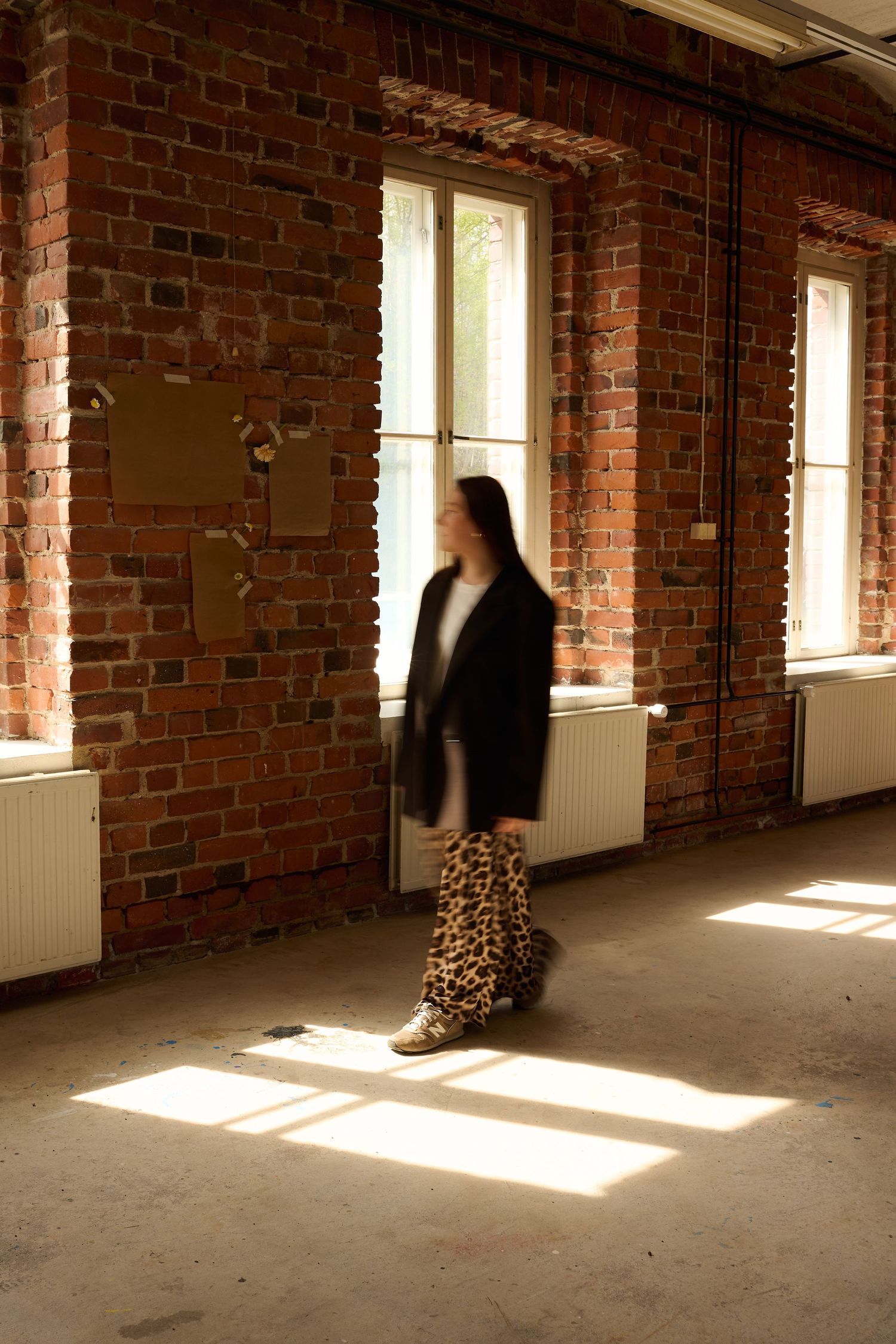 Woman in leopard print pants and black blazer walking in sunlit brick room with large windows.