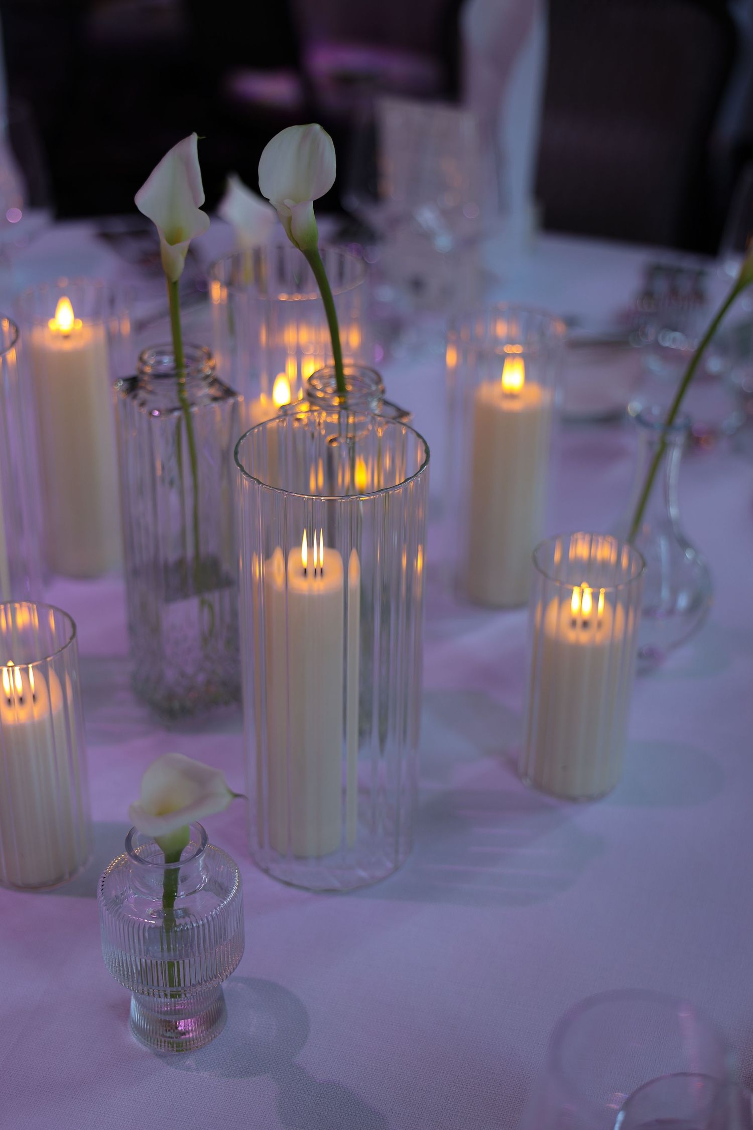 Elegant wedding table centerpiece with lit pillar candles in glass cylinders and white calla lilies in bud vases.