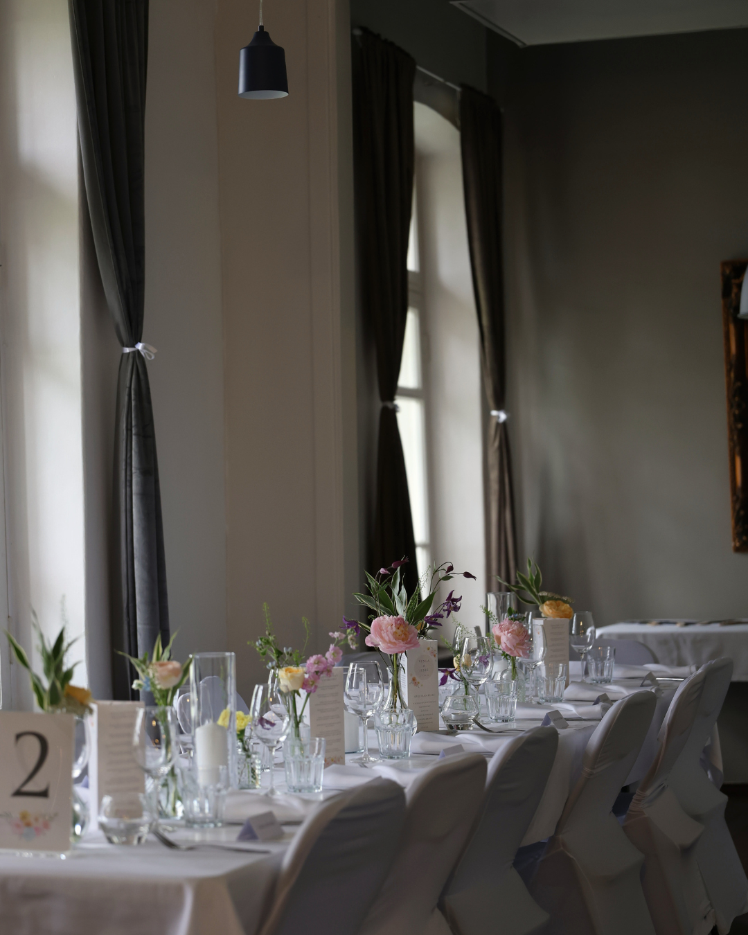 Elegant wedding reception table with white linens, floral centerpieces, and gray curtained windows in a sophisticated venue.