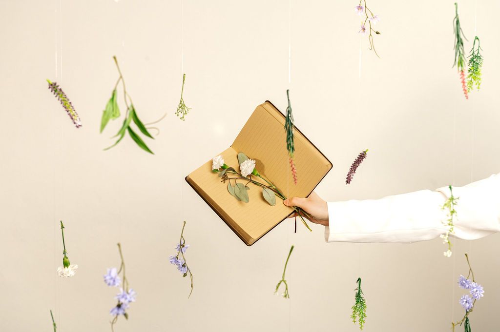 A hand holds a kraft paper journal adorned with a dried flower, surrounded by hanging botanical stems on a neutral background.