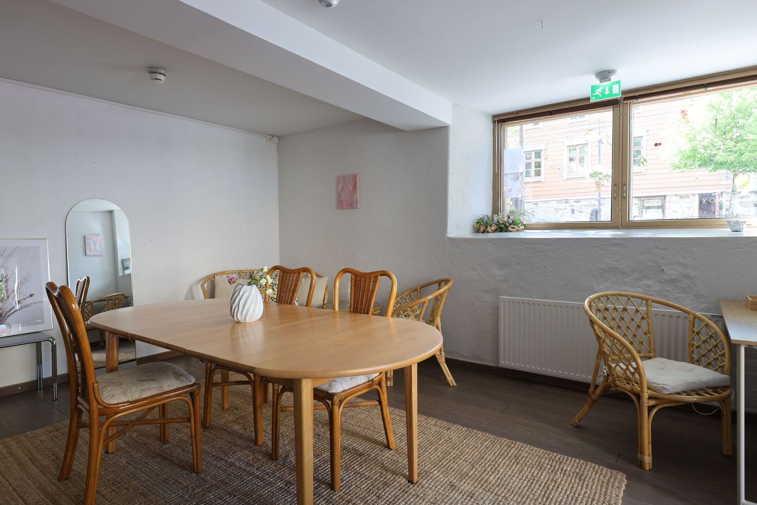 Bright dining room with oval wooden table, rattan chairs, white walls, and a window overlooking a courtyard.