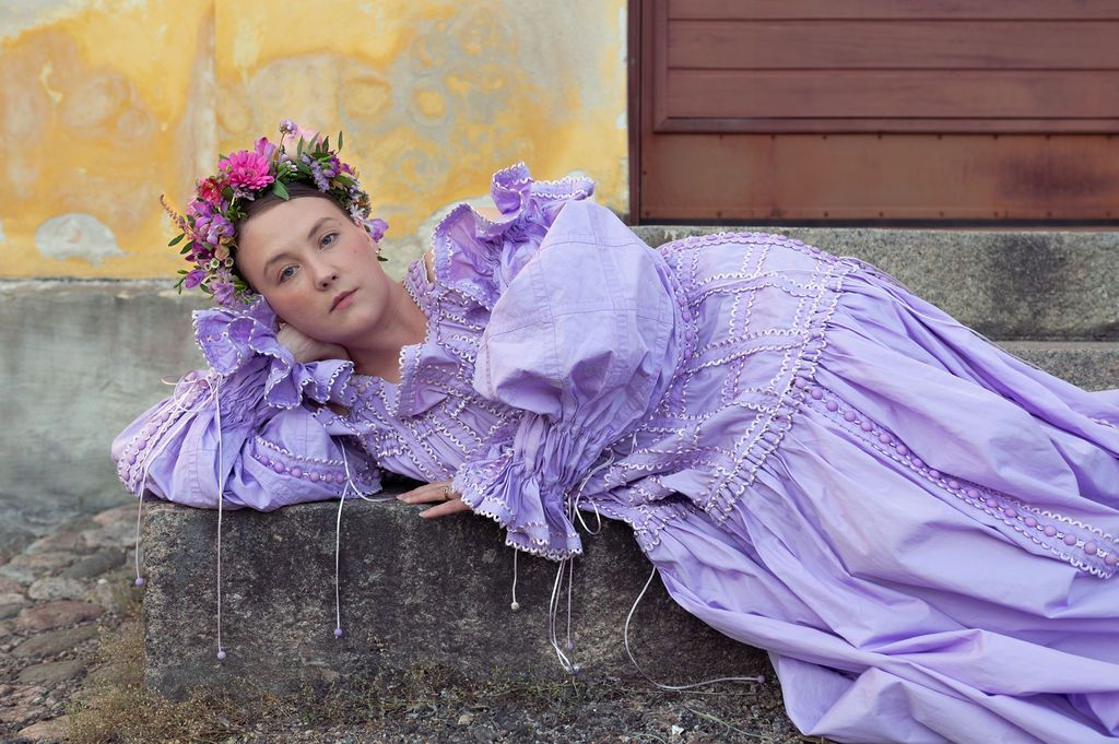 A figure in a flowing lavender dress lies gracefully on rustic stone steps, creating a romantic and ethereal scene.