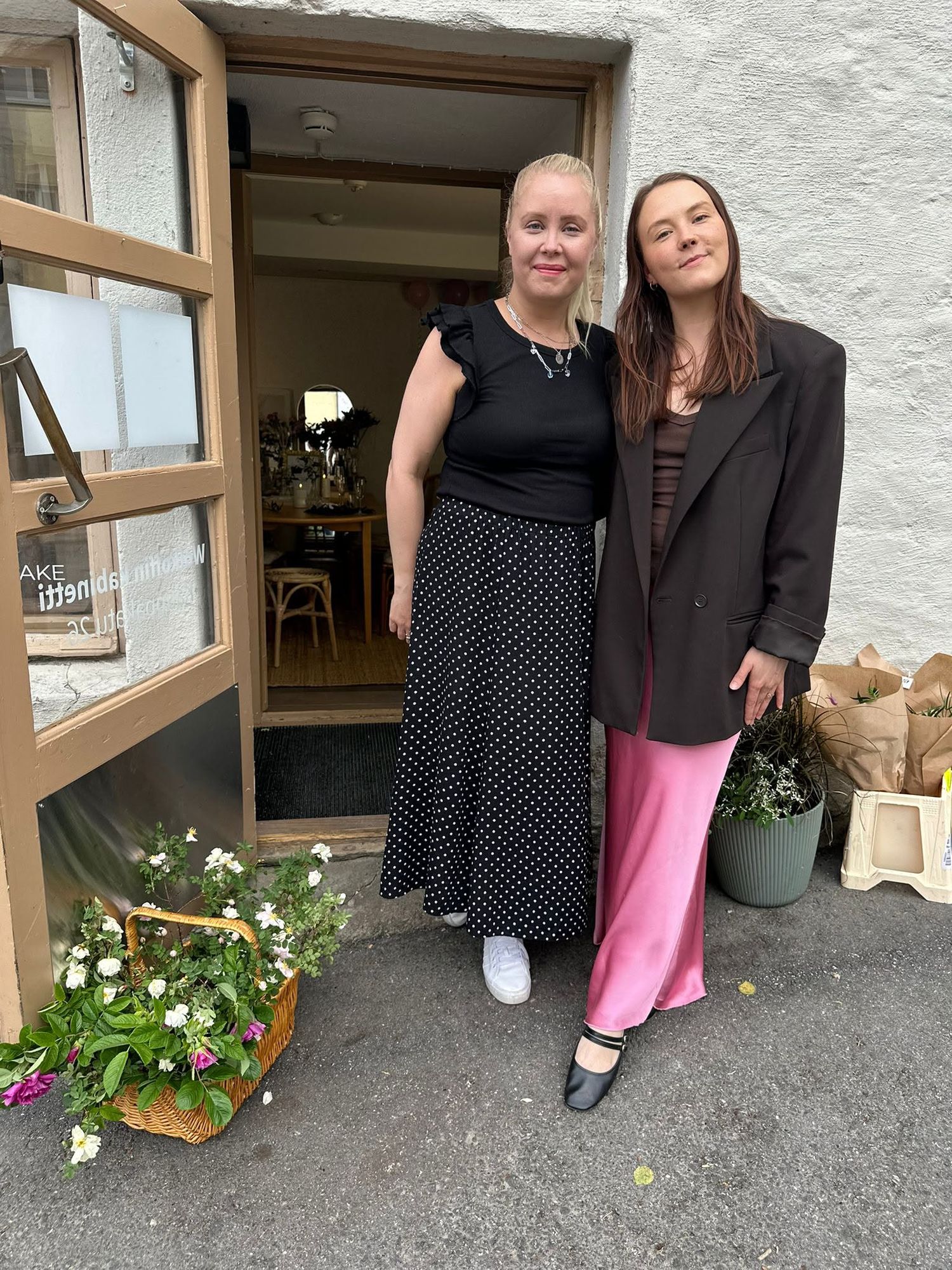 Two women smiling together outside a shop entrance, one in a black polka dot skirt, the other in a pink satin skirt.