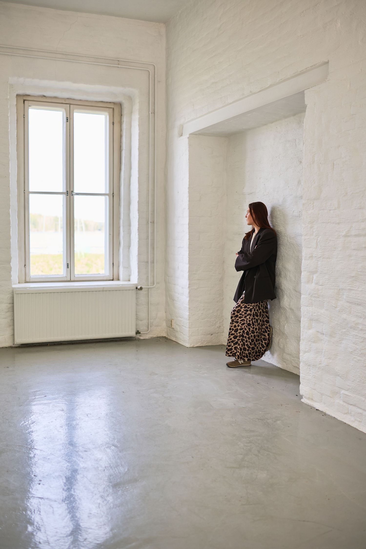 Woman in leopard skirt and blazer leaning against white brick wall in minimalist empty room with large window.