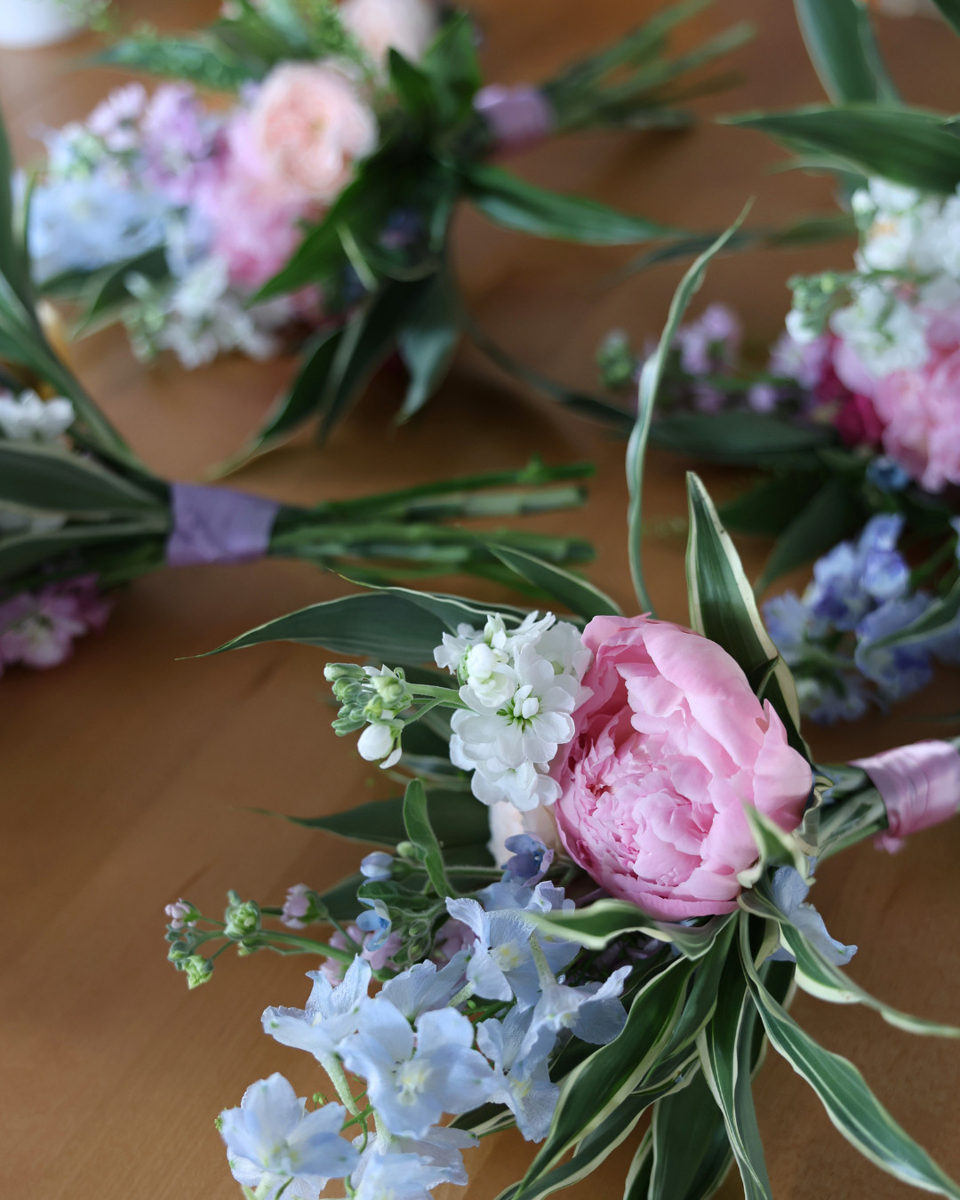 Pink peony boutonnieres with blue delphinium and green foliage arranged on a wooden table for a spring wedding.