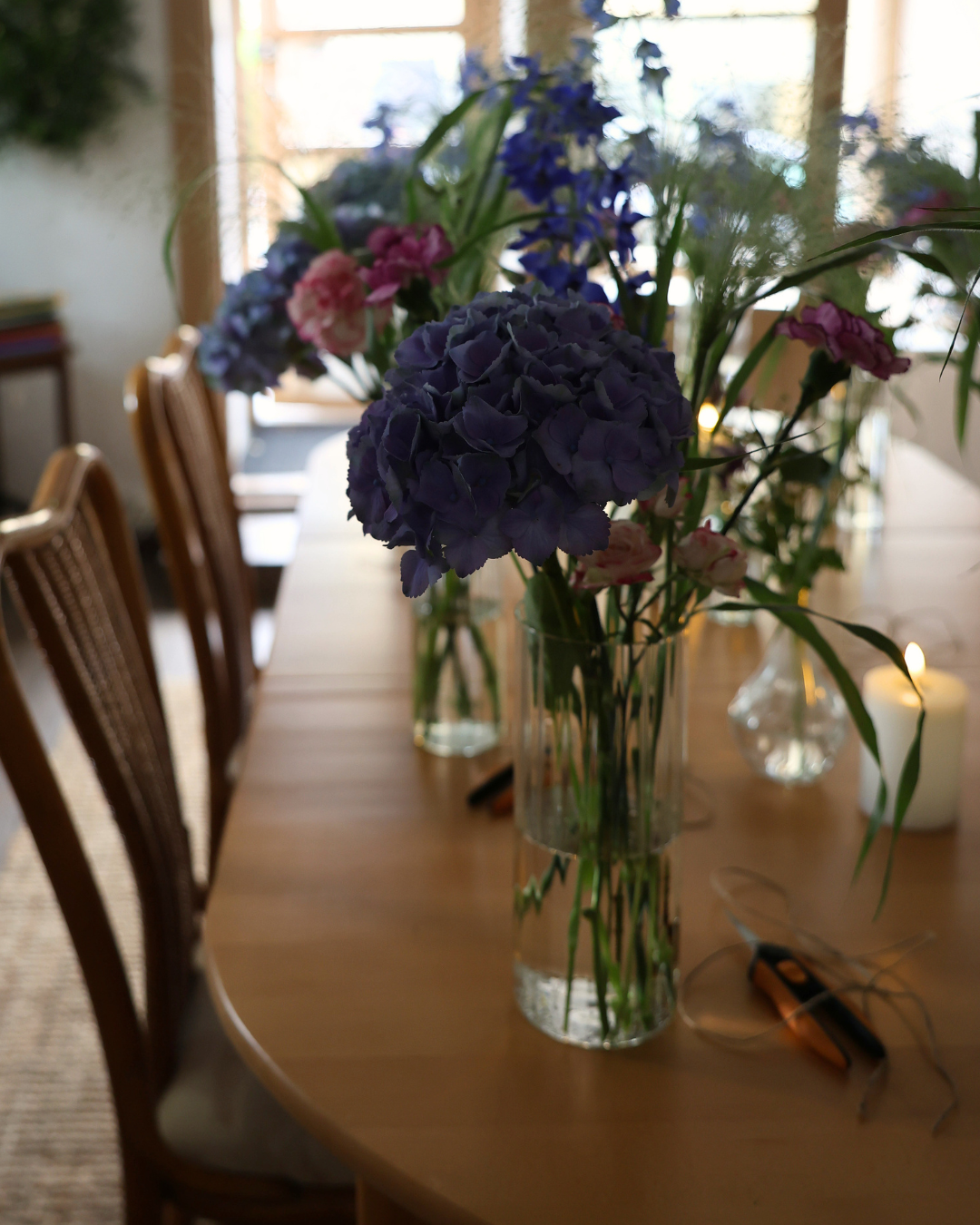 Glass vases with blue hydrangeas and wildflowers arranged on a wooden dining table with candles in a warmly lit room.