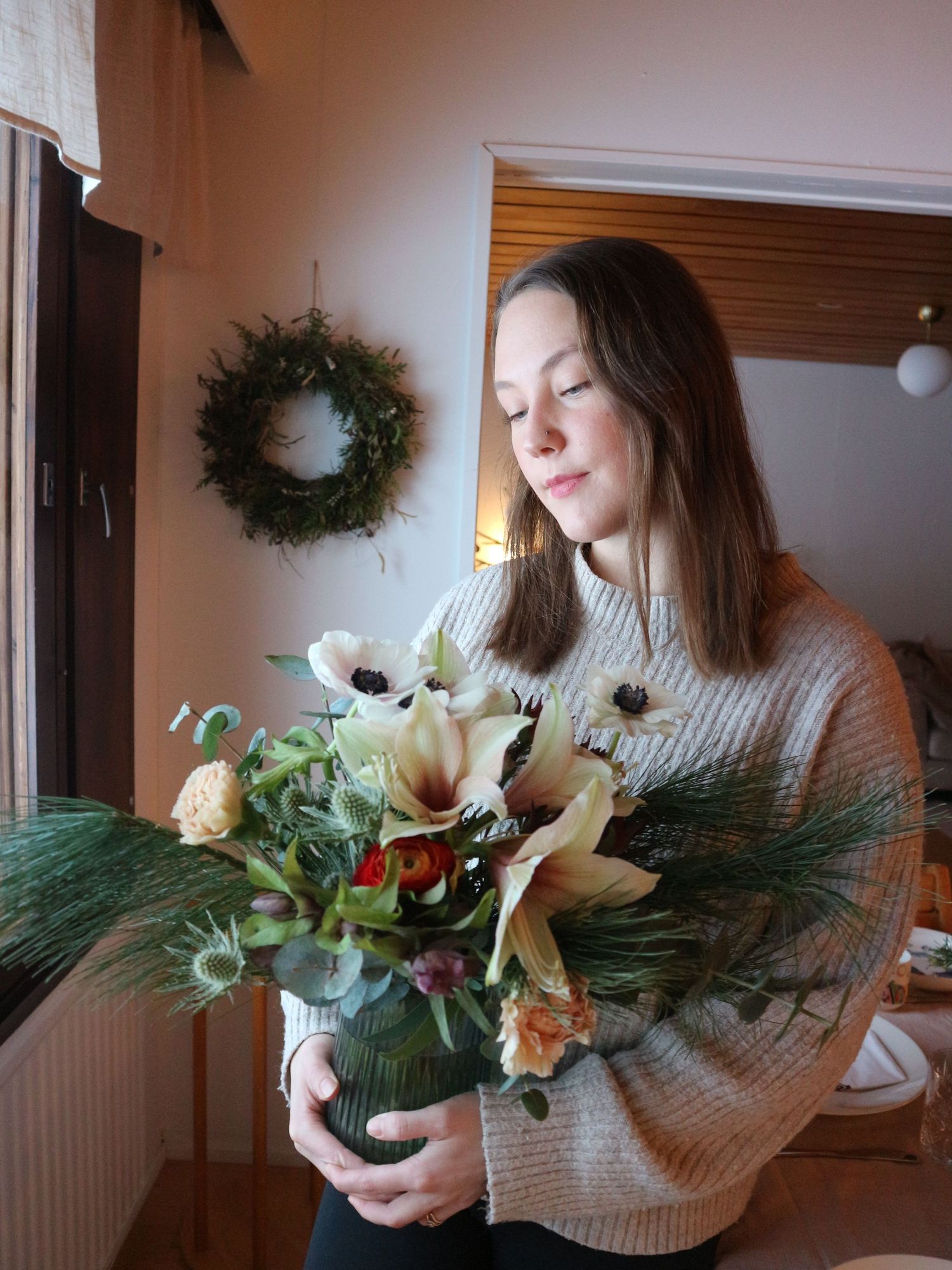 A person in a gray sweater arranges white flowers and evergreen branches in a decorative holiday arrangement.