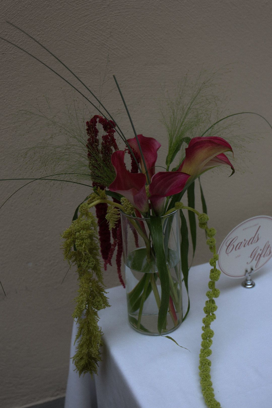 Glass vase with burgundy calla lilies and green amaranthus on a white table next to a cards and gifts sign.
