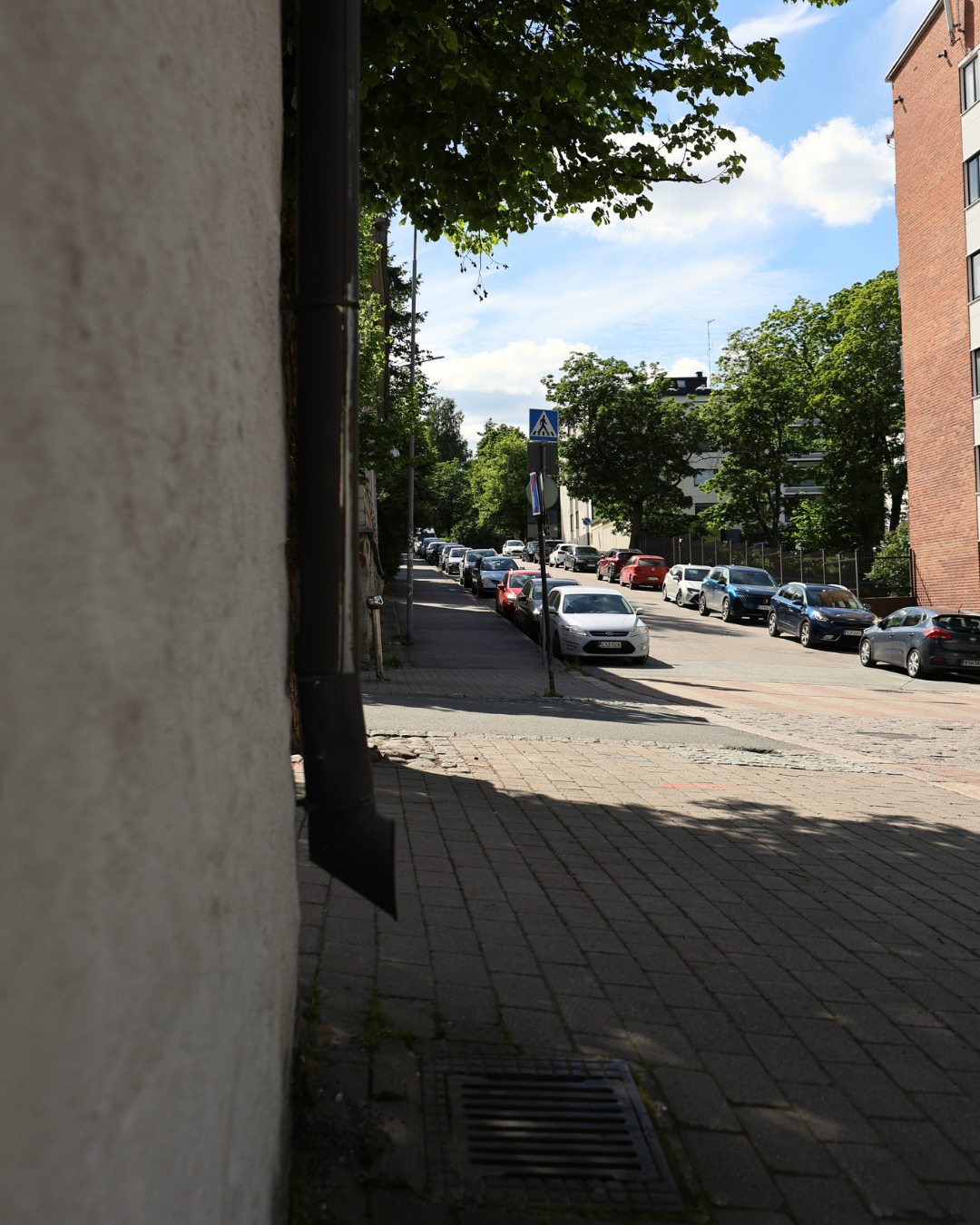A quiet urban street lined with parked cars, brick sidewalks, and lush green trees on a sunny day.