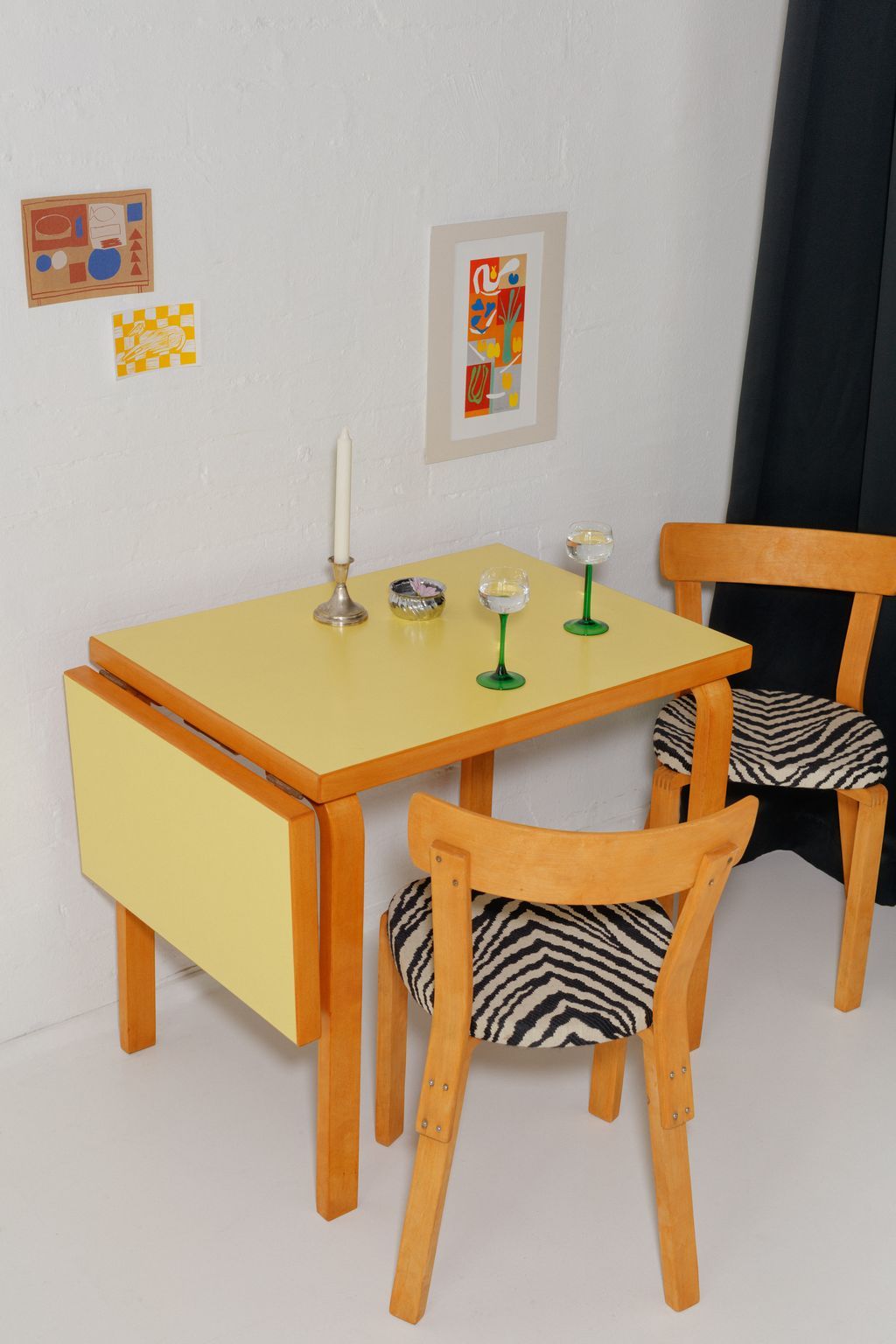 Yellow drop-leaf dining table with wooden chairs featuring zebra print cushions and decorative candle holders on top.