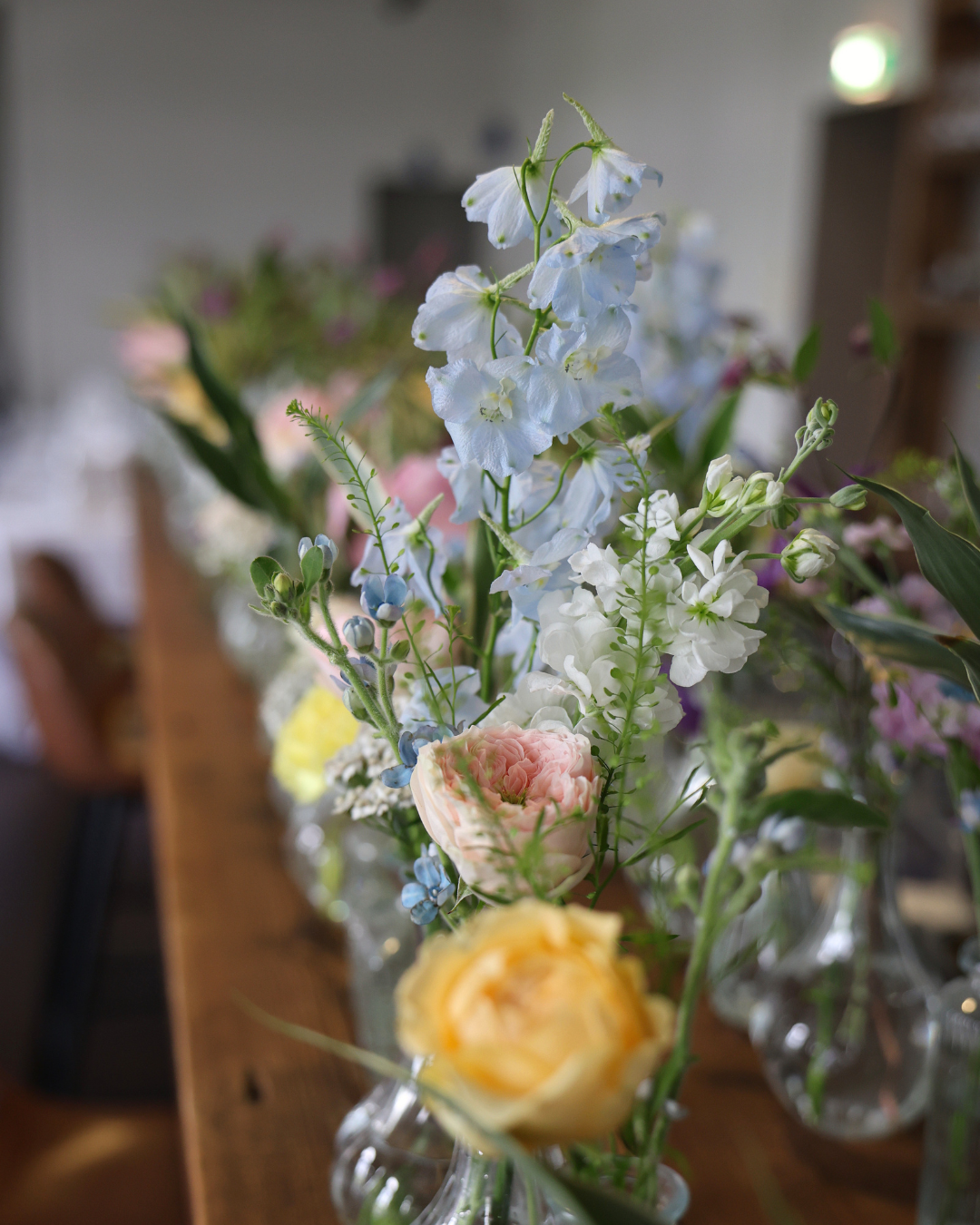 Row of glass bud vases with pastel wildflowers including blue delphiniums and yellow roses on a wooden table.