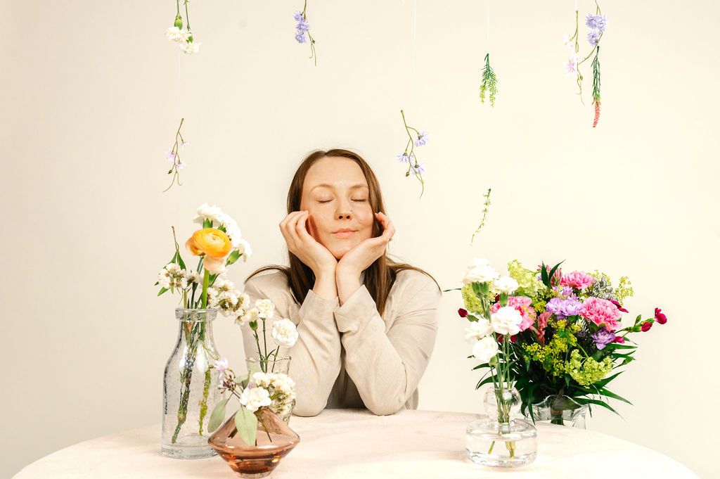 Woman sitting at a table with colorful flower arrangements, eyes closed, hands on cheeks, hanging flowers in background.