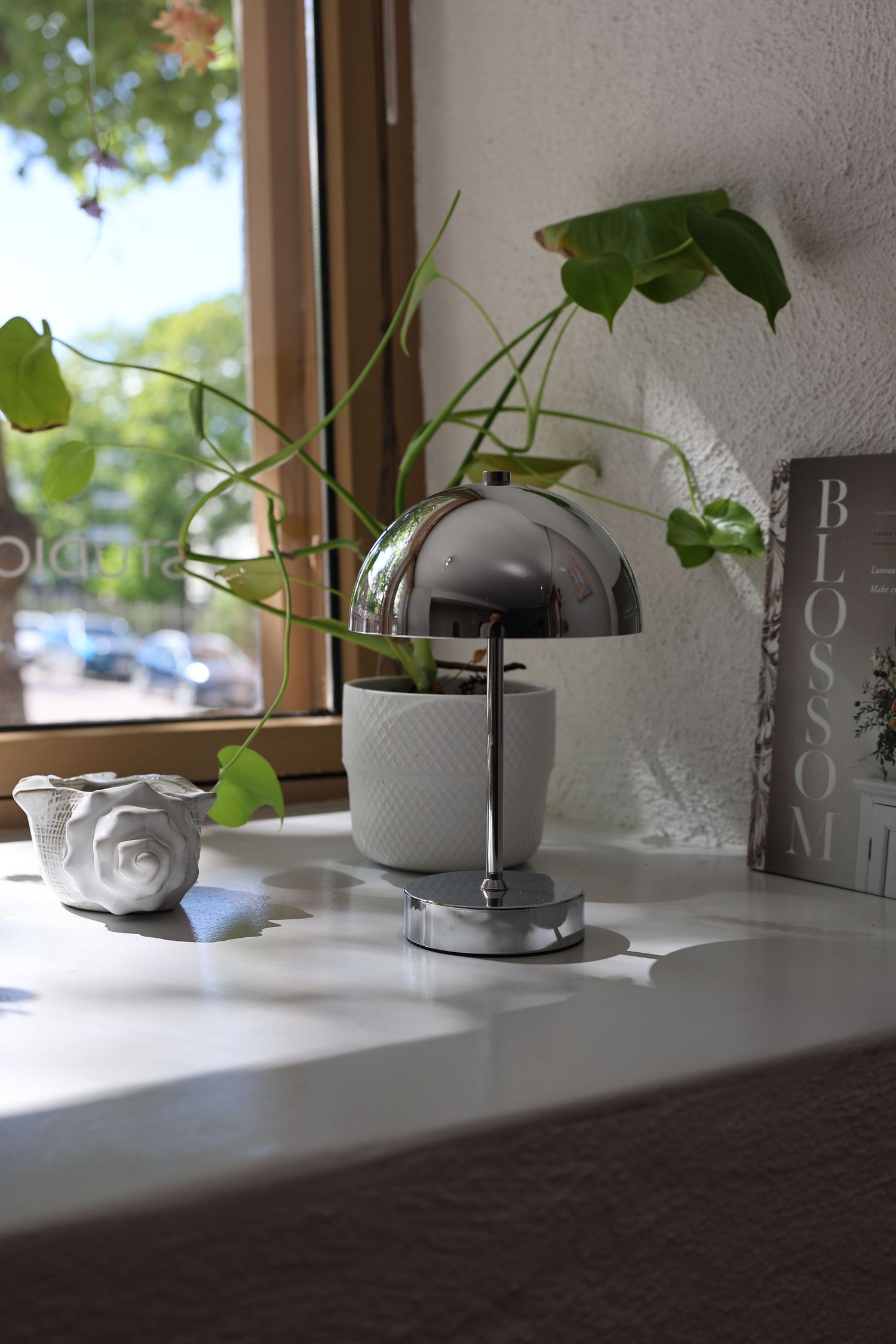 Chrome dome table lamp beside a white ceramic vase with trailing green plants on a white shelf near a sunny window.