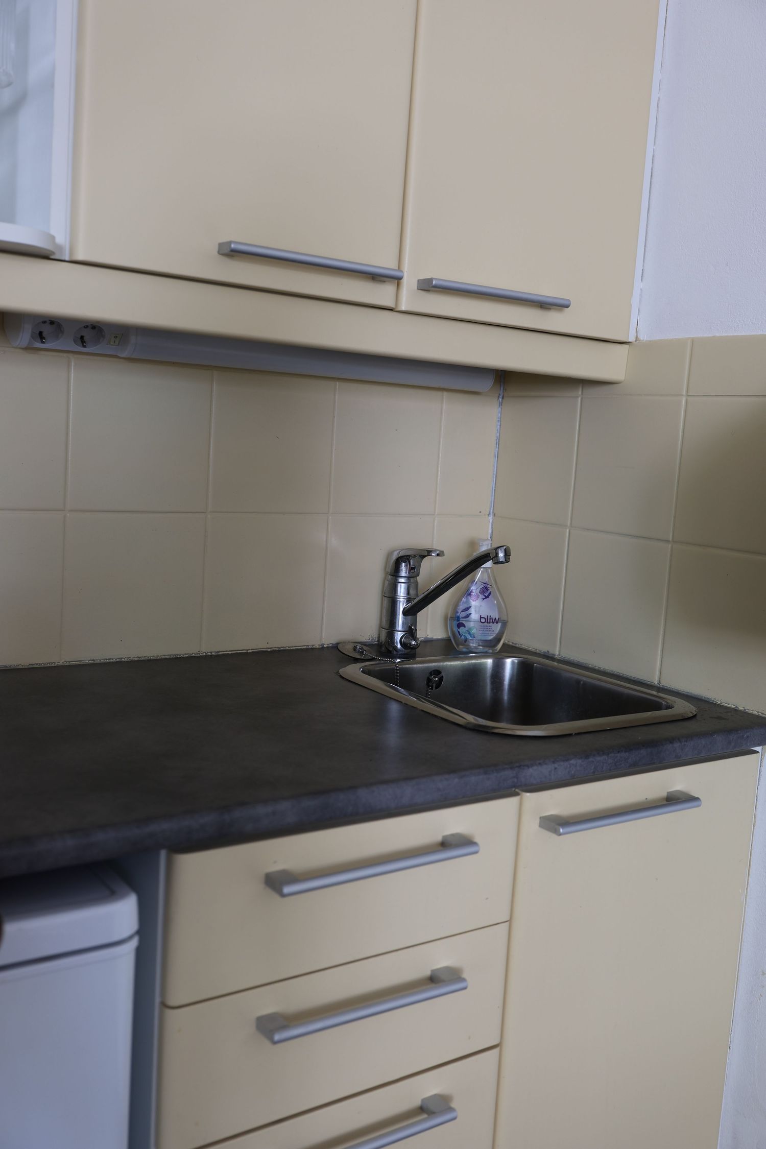 Modern kitchen corner with cream cabinets, dark countertop, stainless steel sink, chrome faucet, and beige tile backsplash.